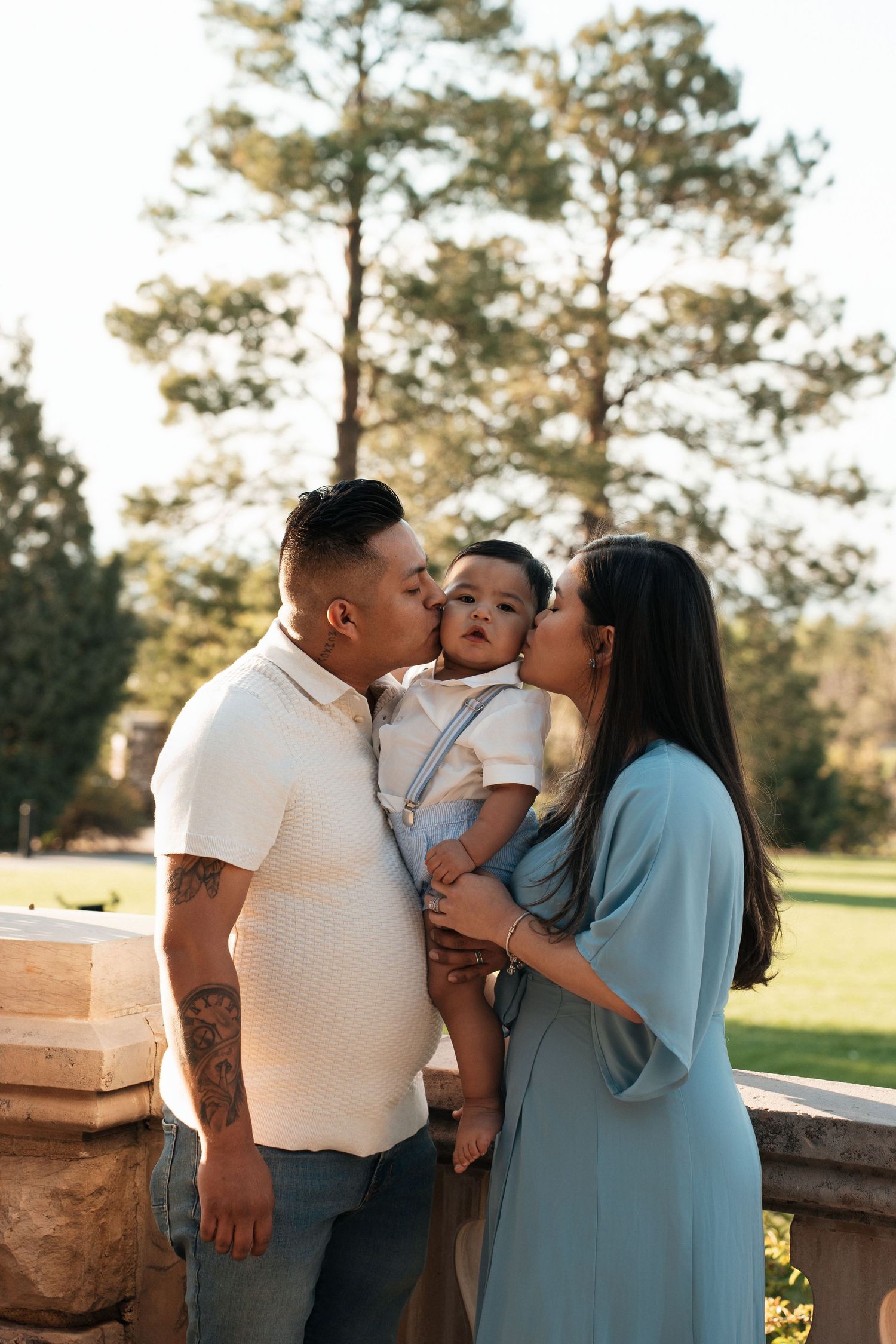 Parents share sweet kisses with their toddler in light-filled outdoor scene with tall pine trees in background.