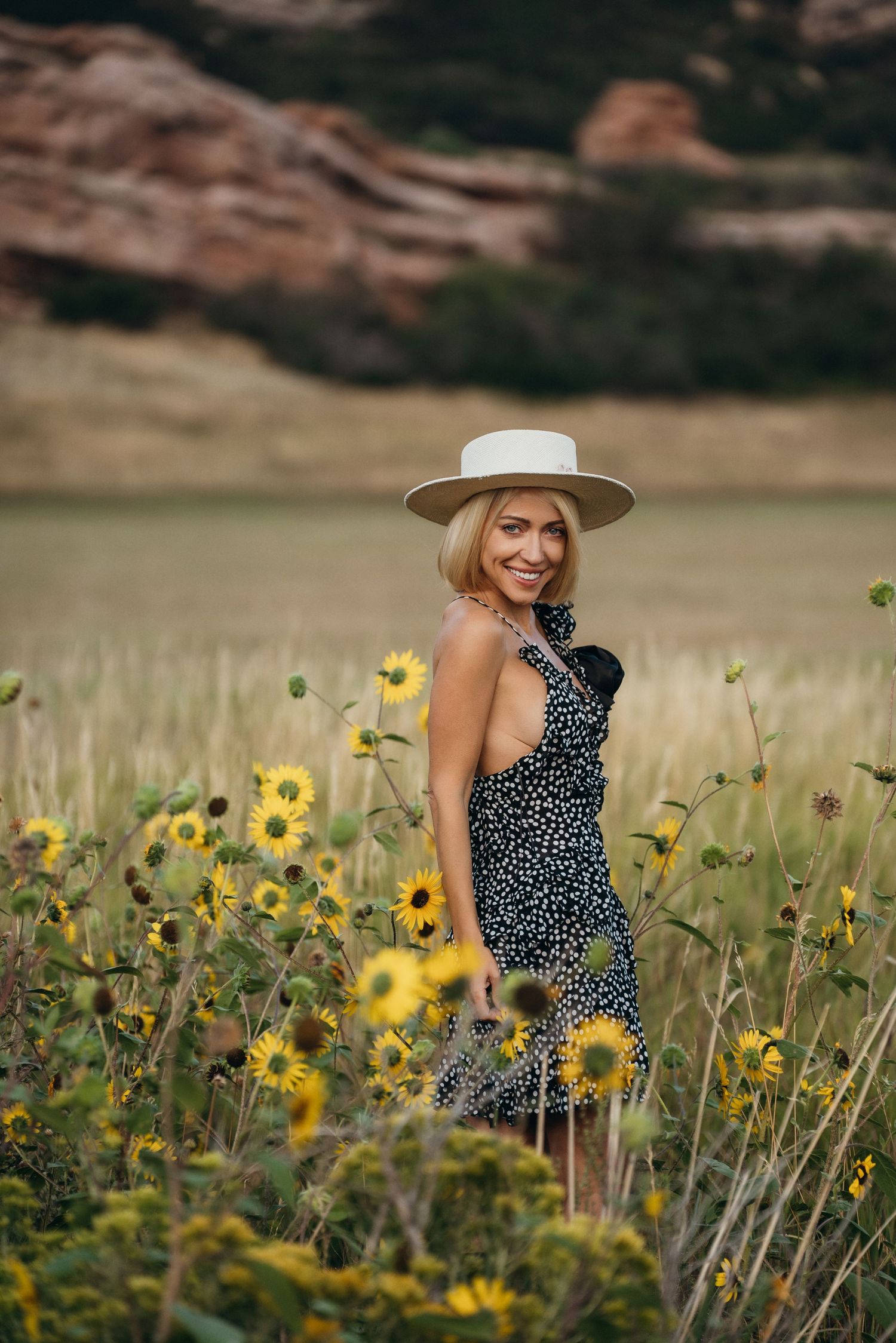A person in a polka dot dress and wide-brimmed hat sits among blooming sunflowers in a scenic field.