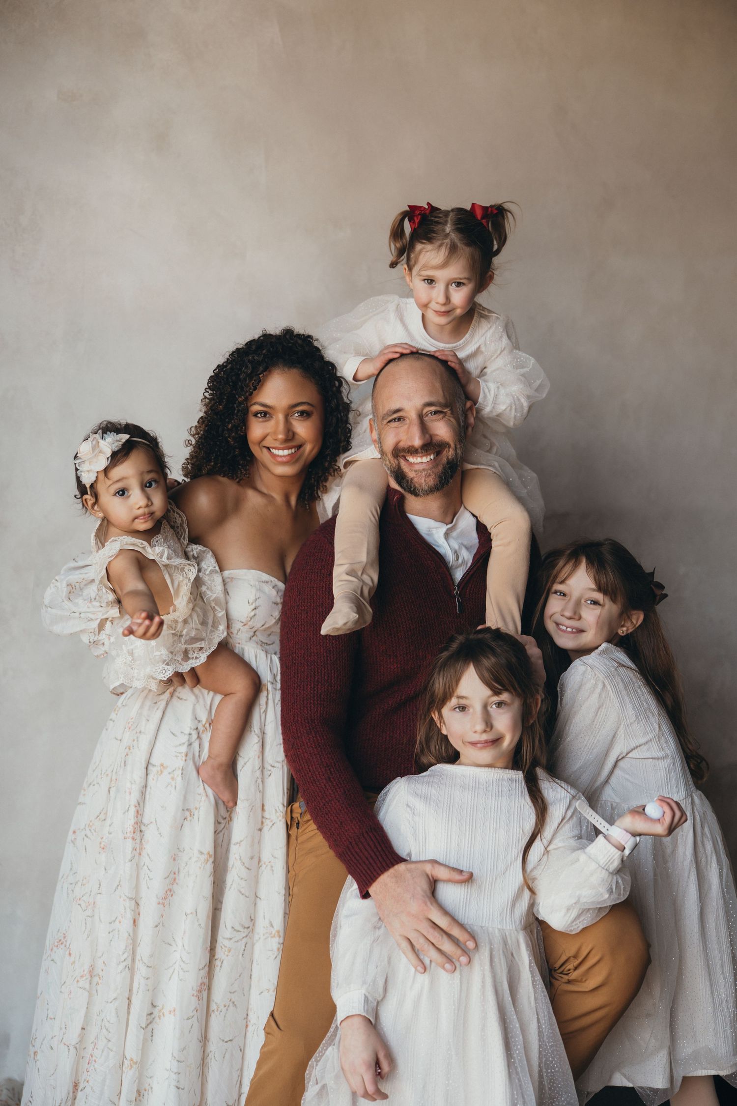 A smiling family of six poses together, with parents holding and surrounded by four young daughters in white dresses.