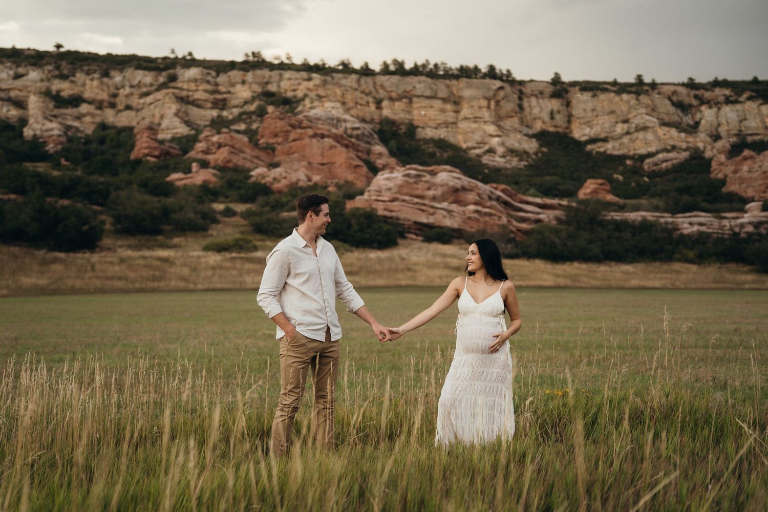 Couple in white outfits walking through a scenic field with dramatic rock formations in the background at sunset.