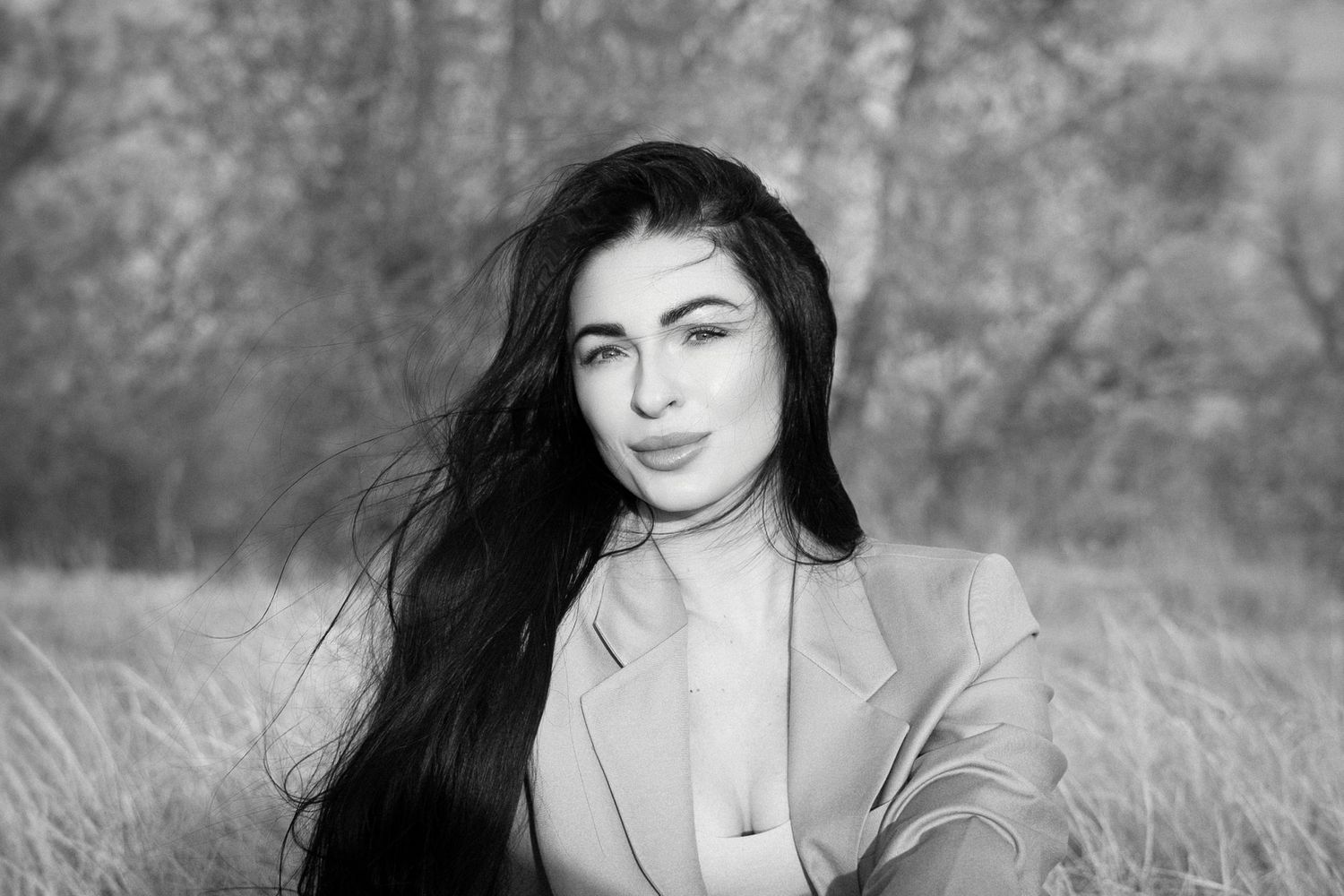 Black and white portrait of woman in beige blazer with long dark hair outdoors in nature setting.