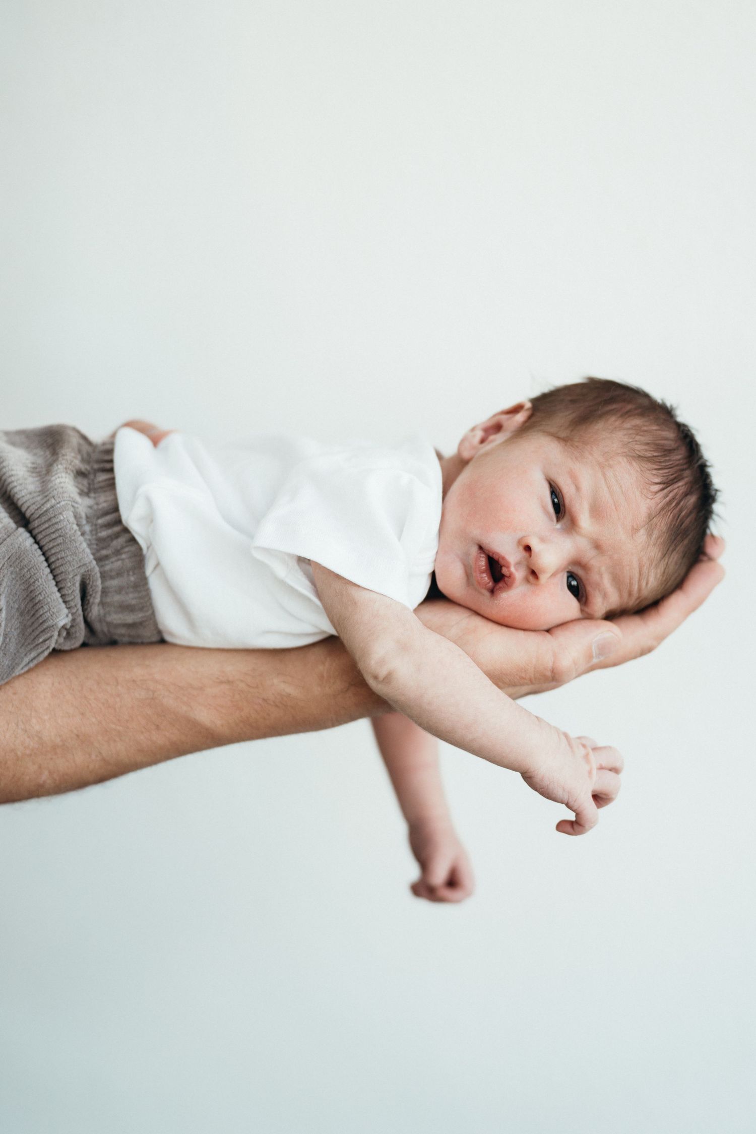 A small baby wearing a white shirt and gray pants lies playfully across an adult's arm against a light background.