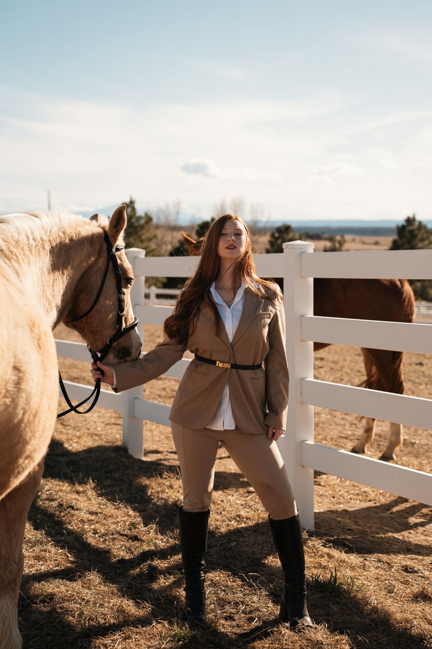 An equestrian in riding attire stands next to a white horse by a wooden fence at sunset.