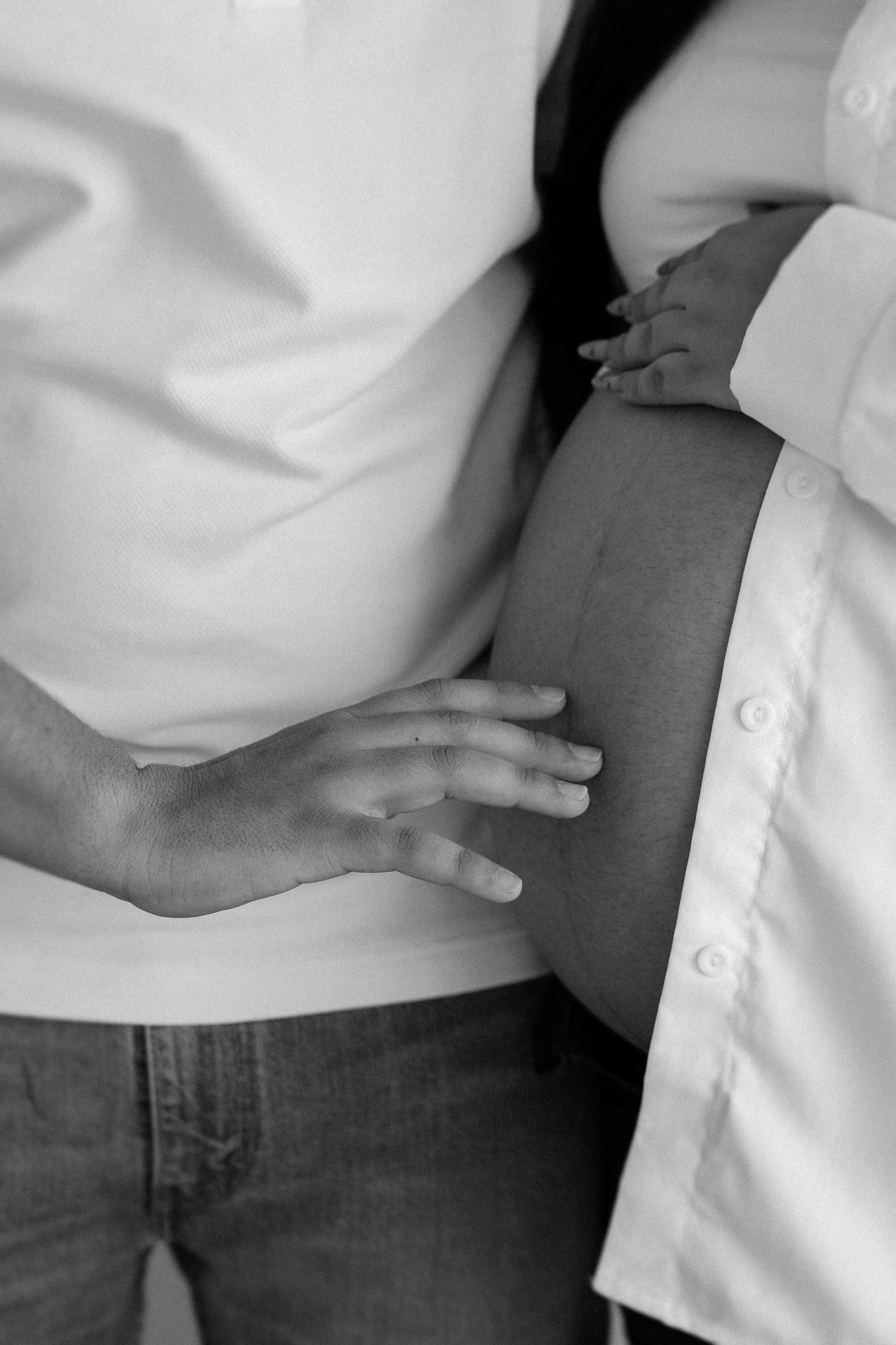 A black and white close-up photo showing hands gently resting on a pregnant belly, wearing denim and a white shirt.