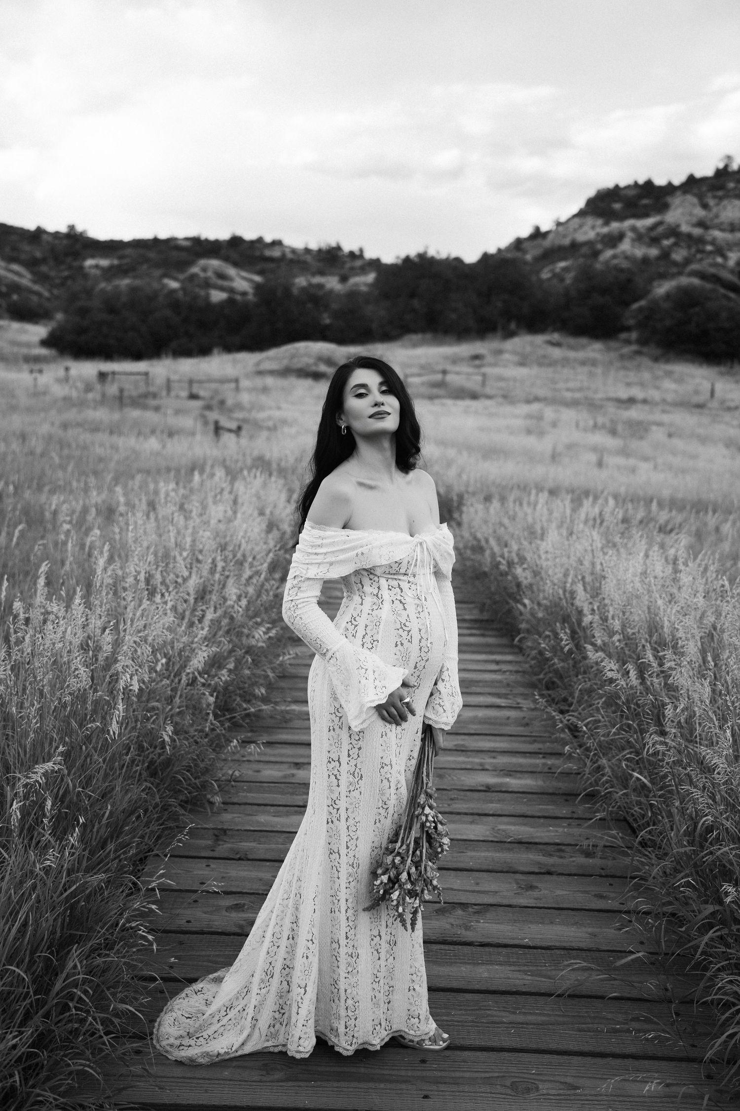 A black and white photograph of a lace wedding dress being modeled on a wooden boardwalk through a grassy field.