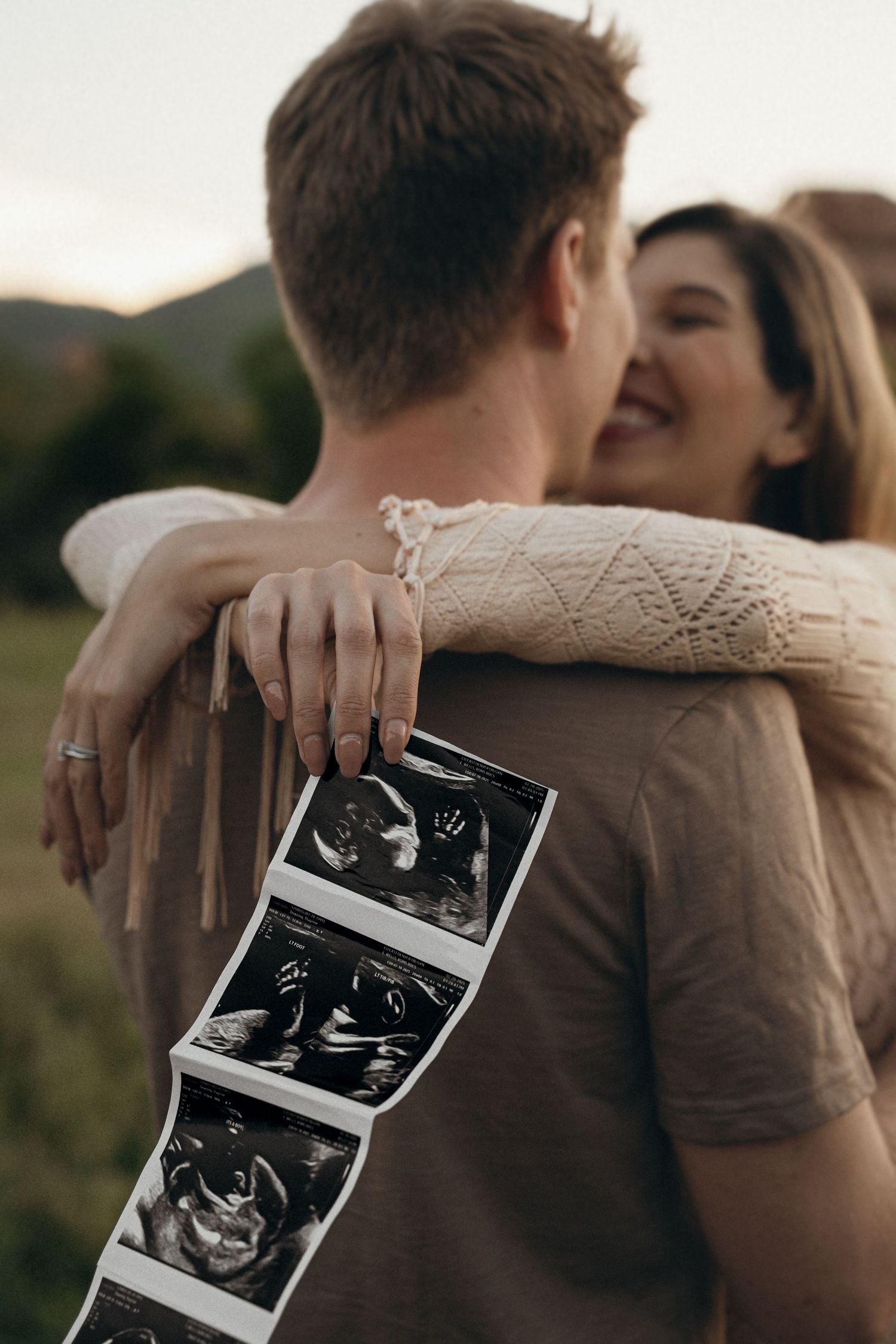 A couple embraces outdoors while holding ultrasound photos, celebrating their pregnancy announcement in warm sunlight.
