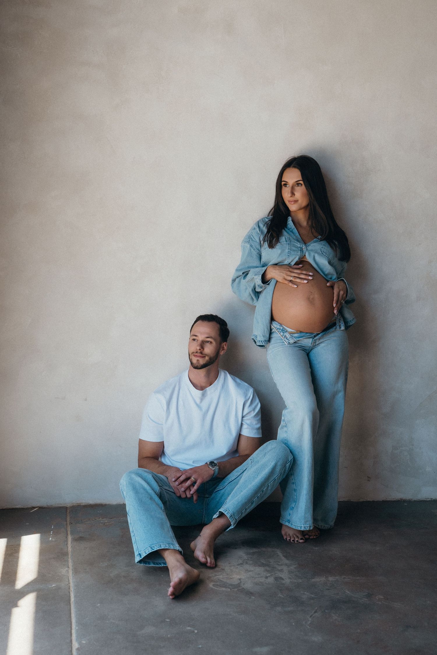 A pregnant woman in denim holds her belly while a man sits on the floor beside her against a gray wall.