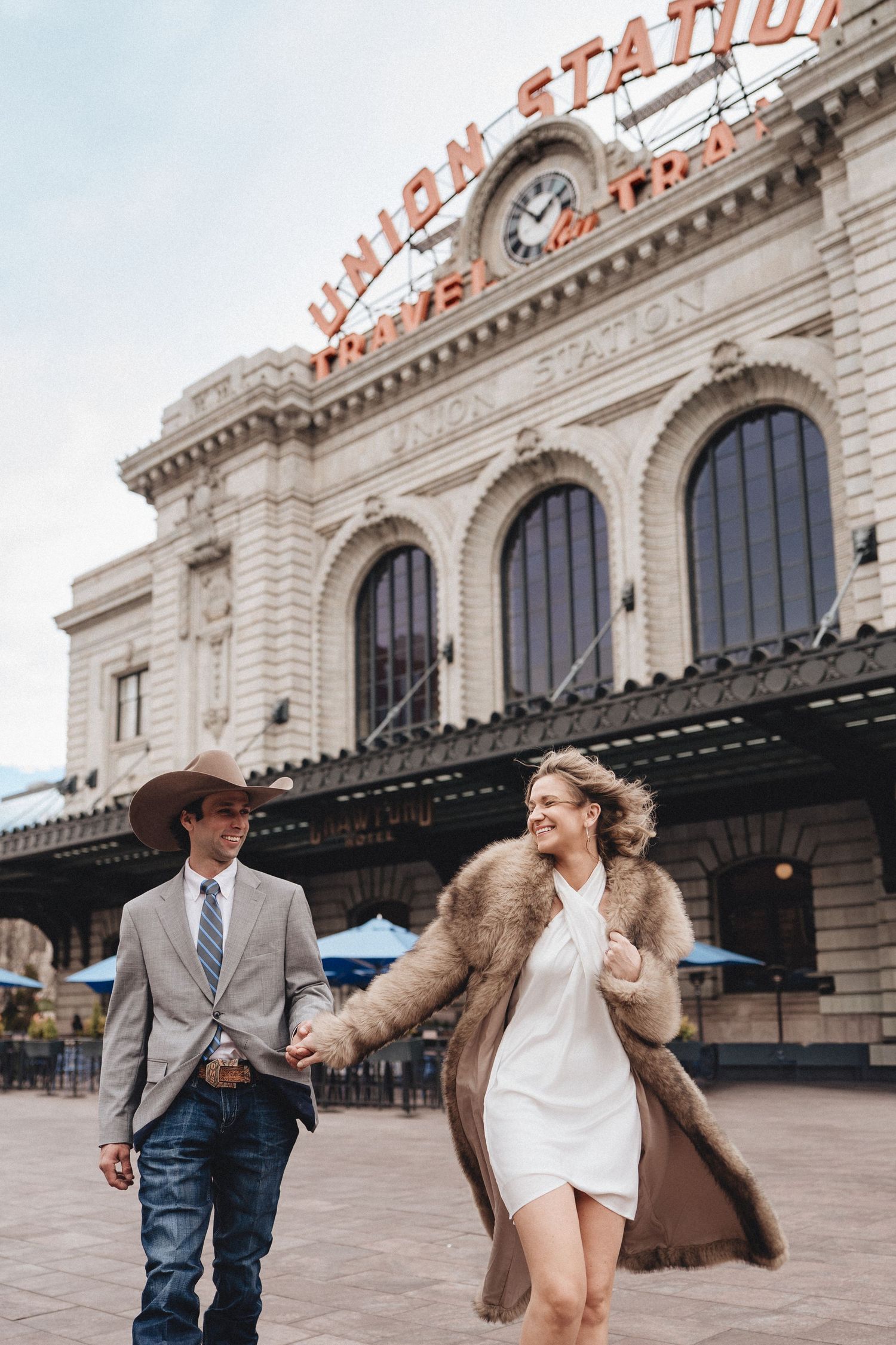 A couple in stylish attire walking hand in hand in front of Denver's historic Union Station building.
