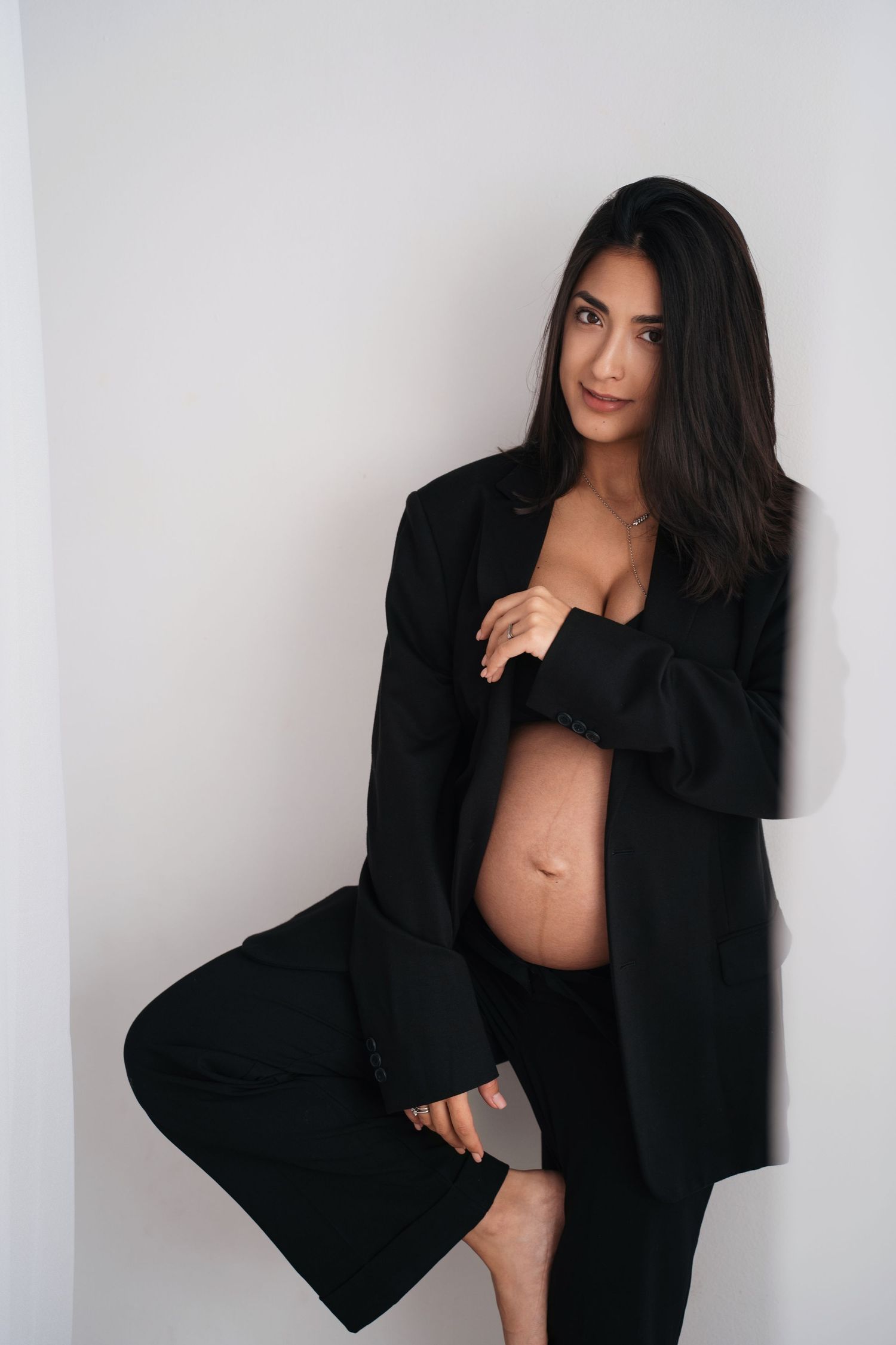 Studio maternity portrait with woman in black blazer against white backdrop.