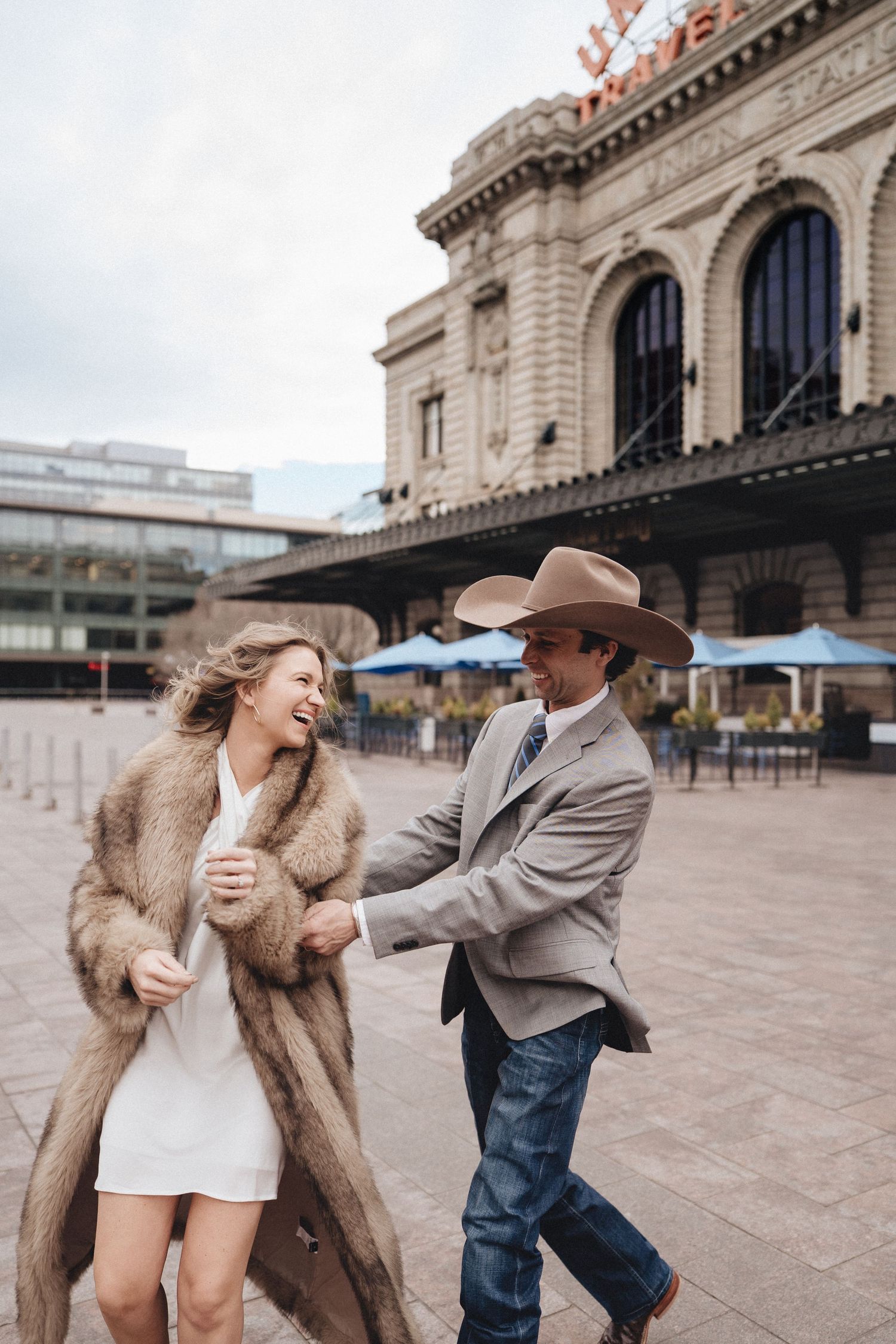 A couple dances playfully in front of a historic train station building, with the woman wearing a fur coat.