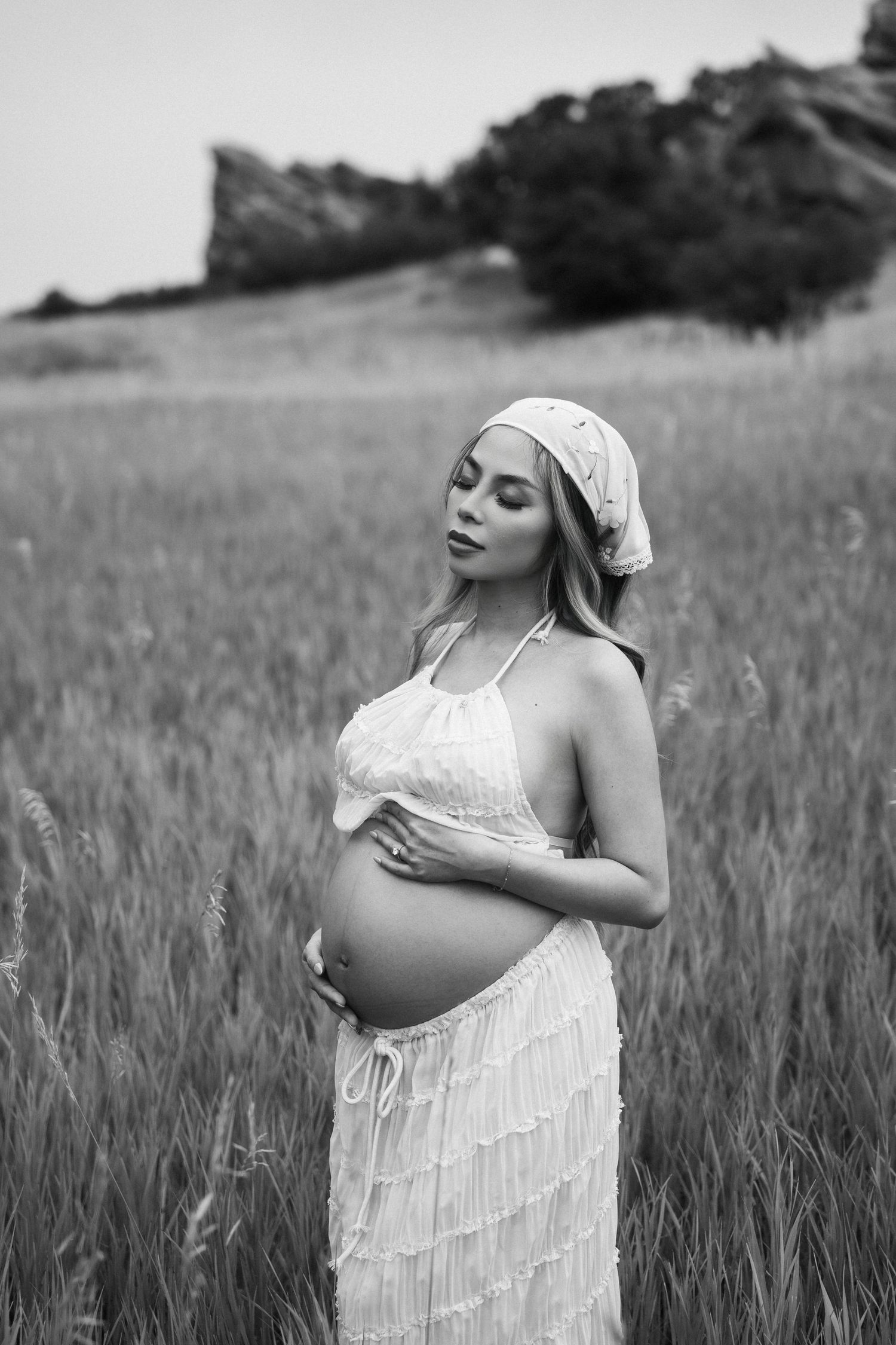 A pregnant woman in a white dress and headscarf stands thoughtfully in a wheat field in black and white photography.