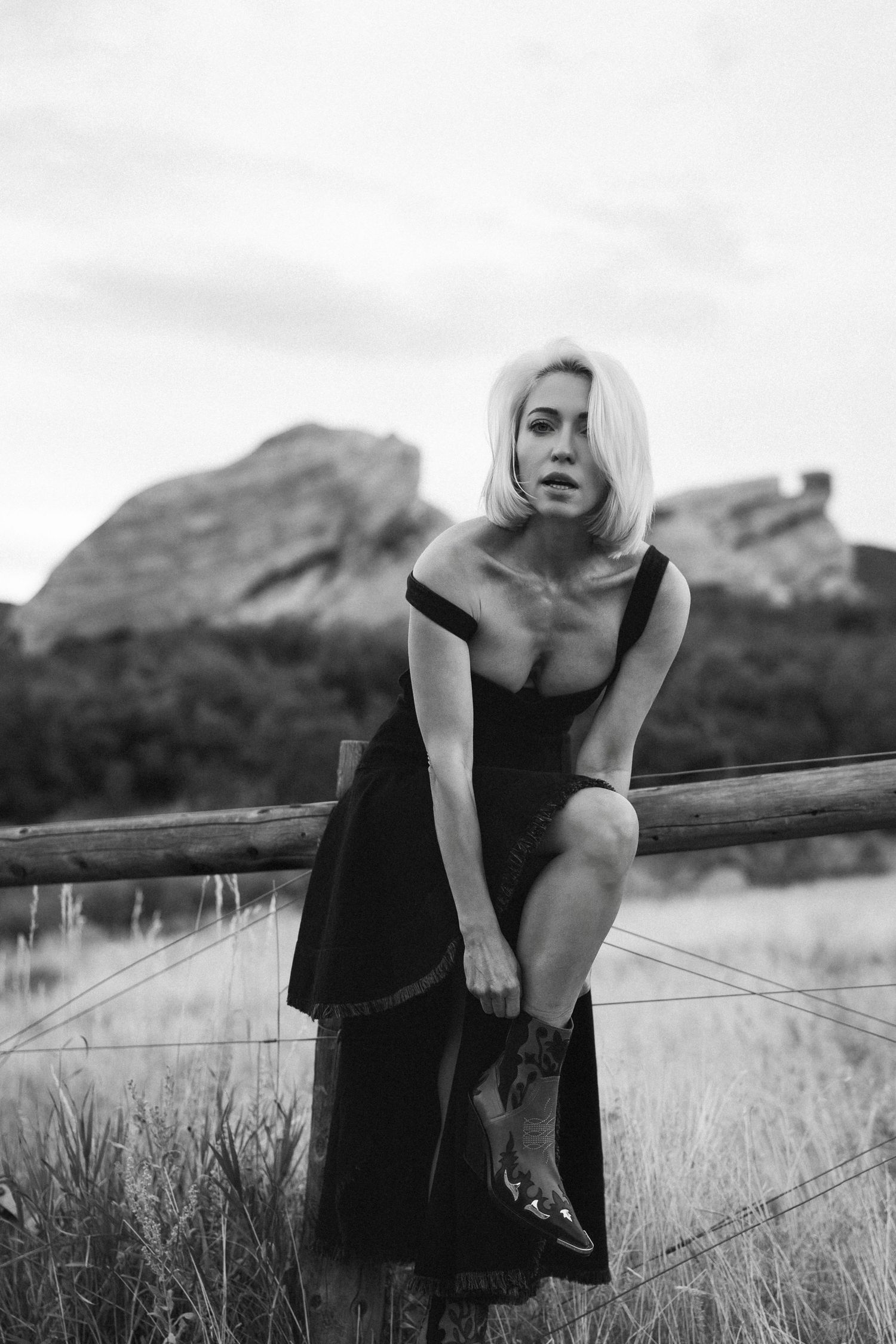 Black and white fashion editorial series showing a model in a dark dress posing in a grassy field with mountains in background.