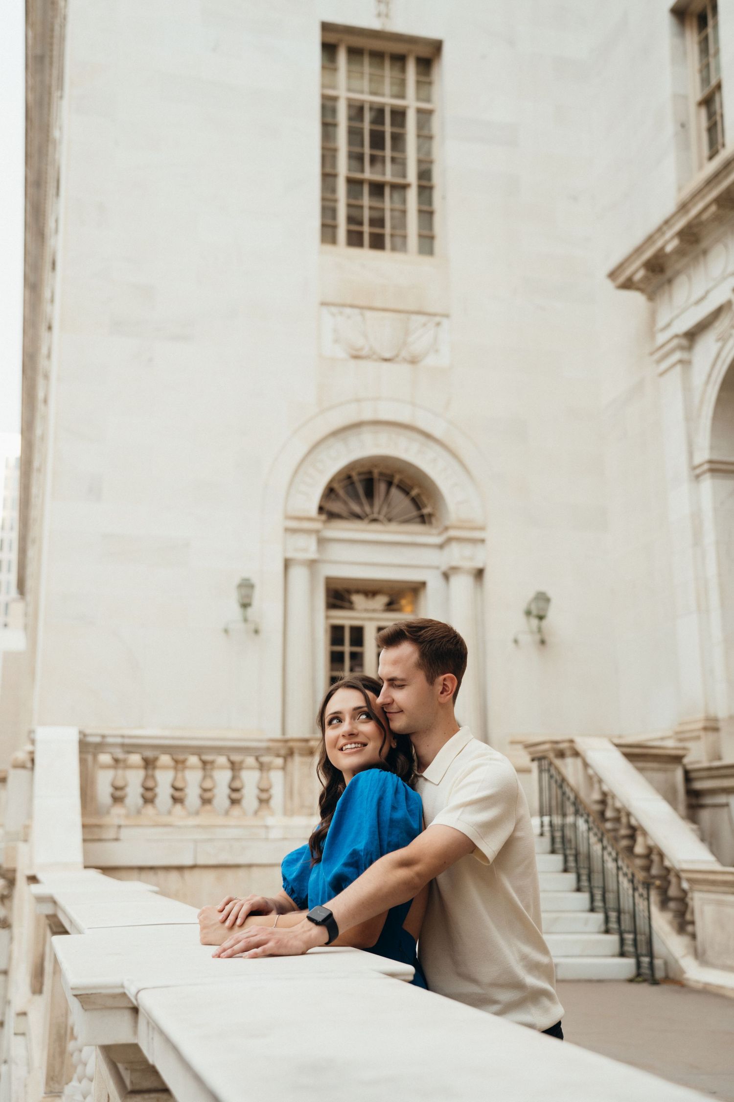 A couple shares a romantic moment leaning against a white stone balcony at an elegant historic building with classical architecture.