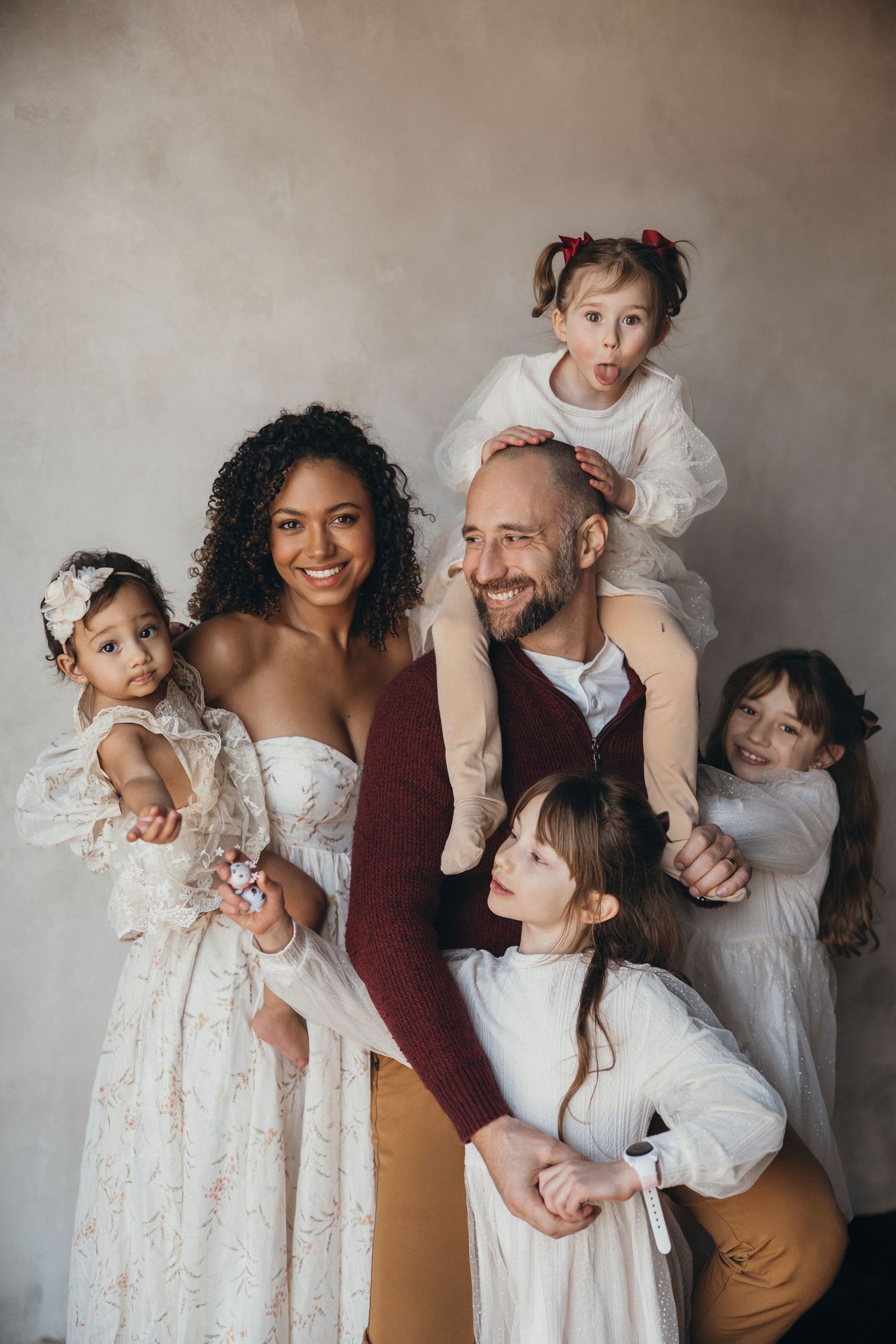 A family wearing white and earth-toned clothing poses playfully together in a series of candid studio portraits.