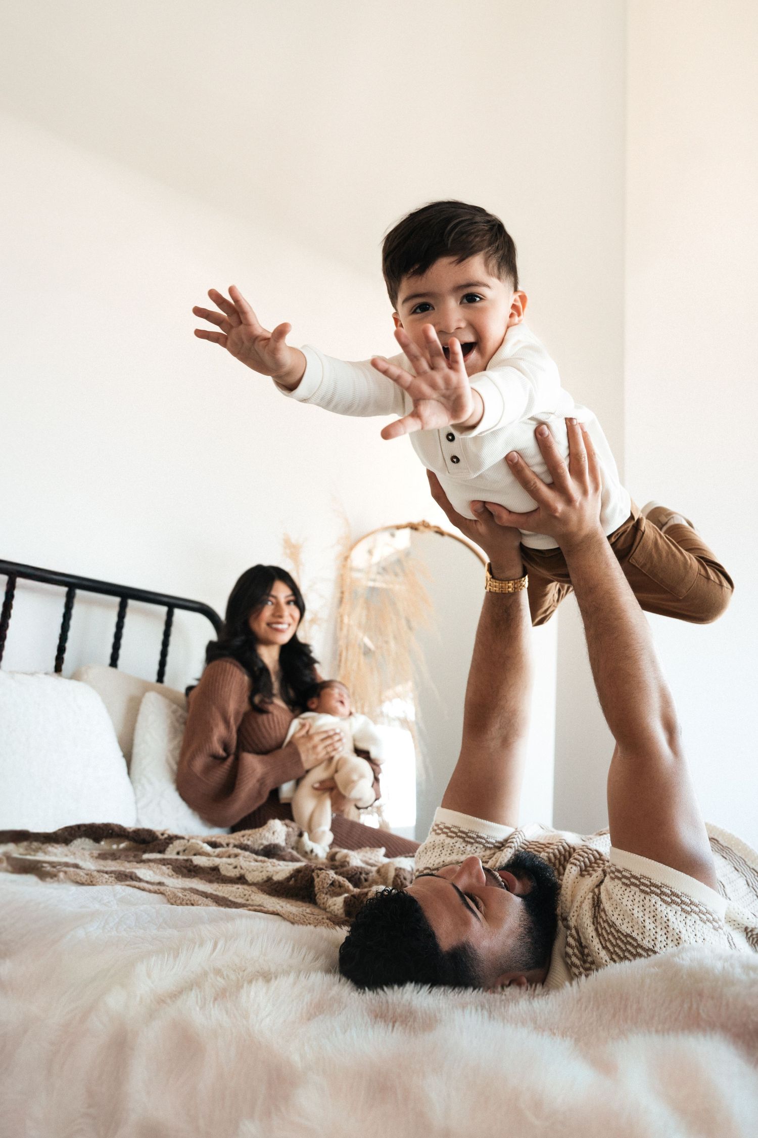 Family enjoying playful moment together on bed as toddler gets lifted high in the air during morning playtime.