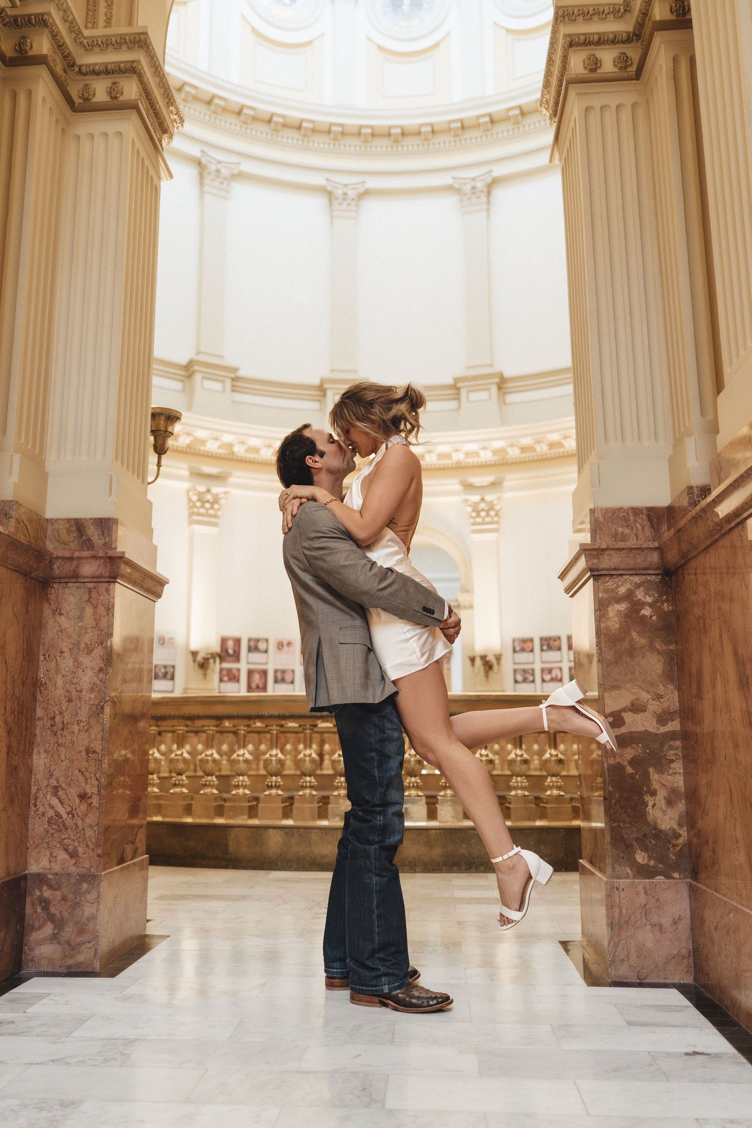 Romantic couple embraces in a grand marble rotunda with ornate architectural details and columns.