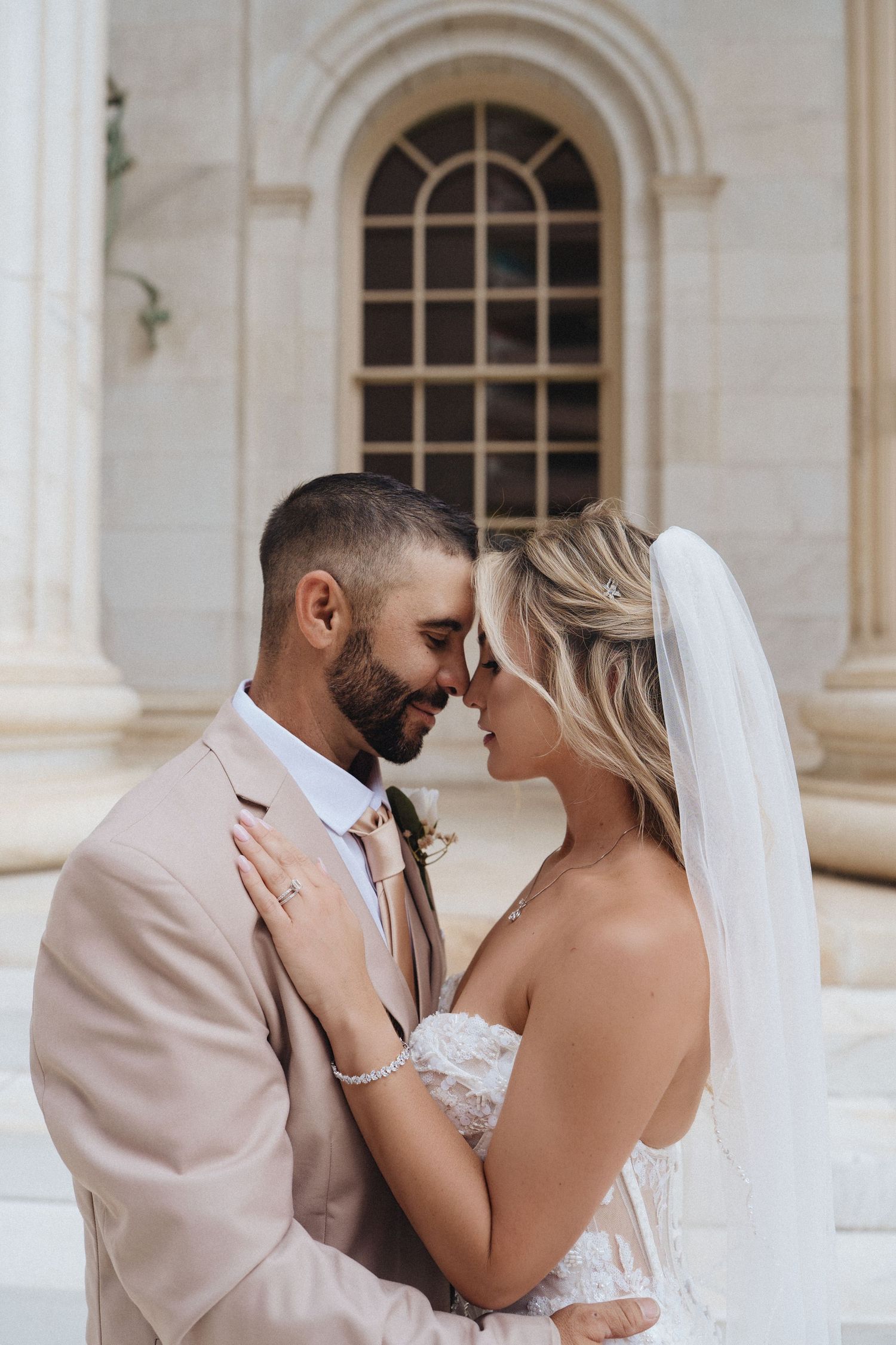 Bride and groom in romantic forehead touch pose, groom in blush suit, bride in lace dress with veil, elegant stone building backdrop.