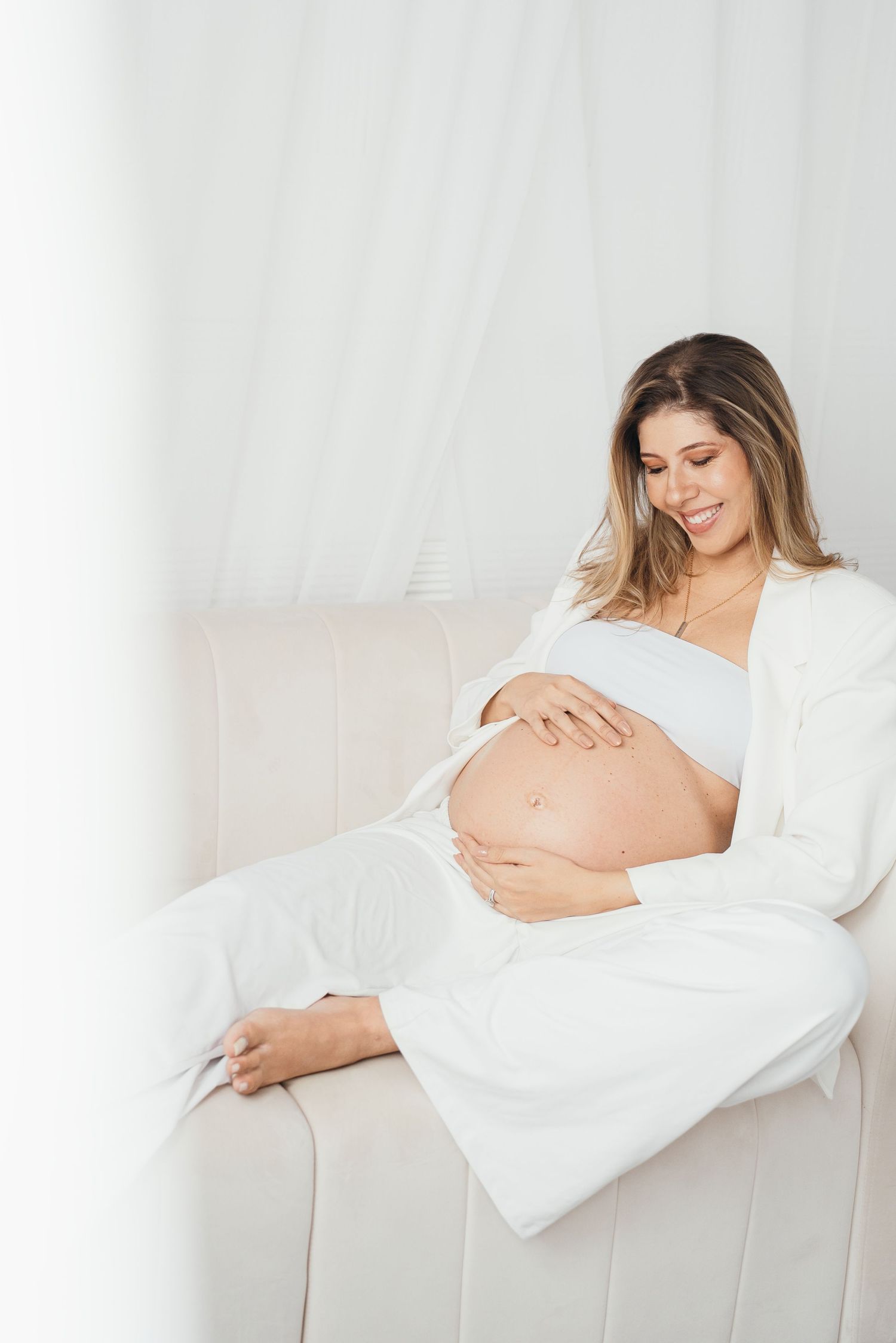 A pregnant woman in white clothing sits peacefully on a cream-colored couch while cradling her baby bump.