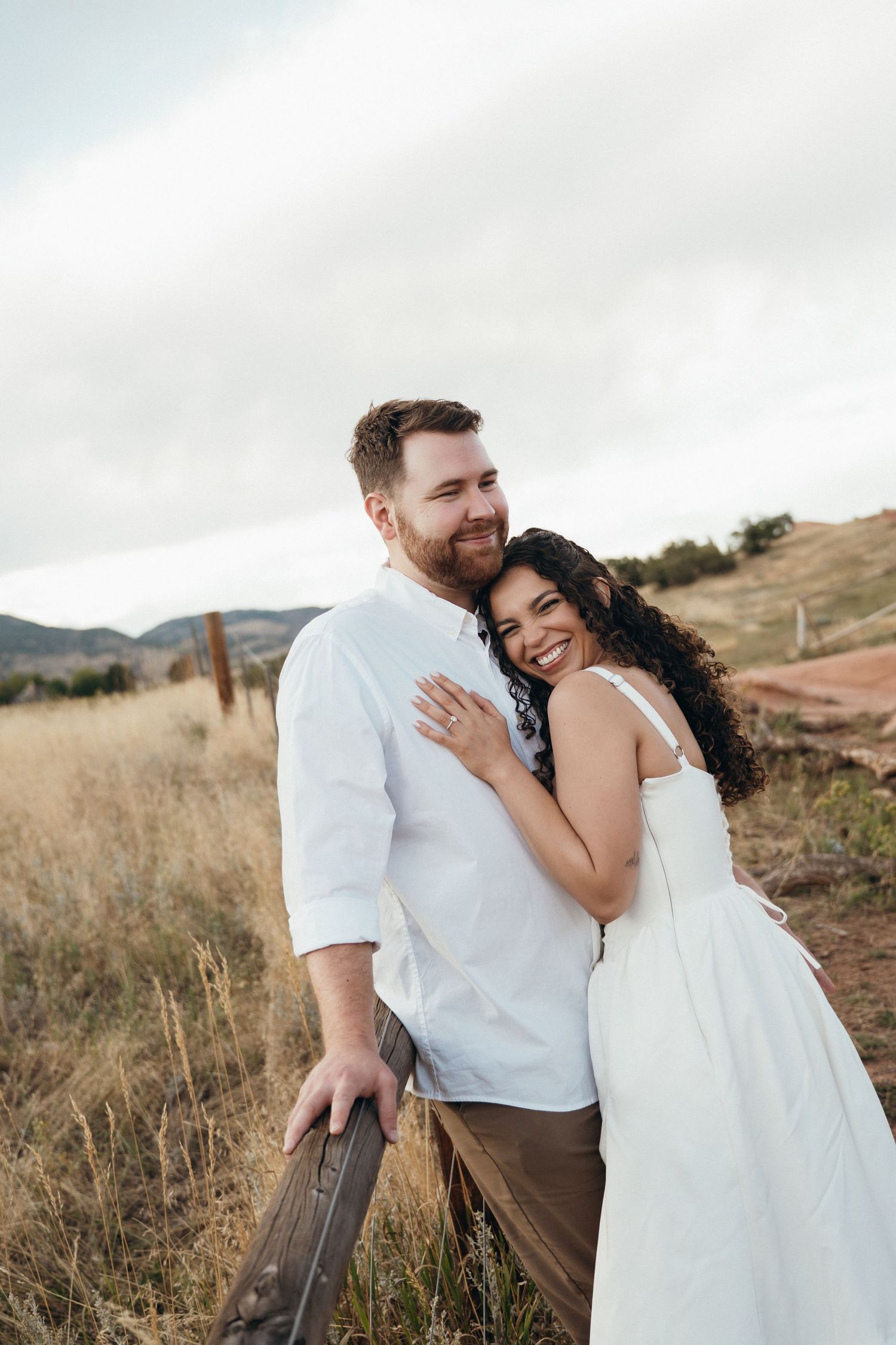 A couple shares an intimate moment in a rustic outdoor setting with mountains and tall grass in the background.