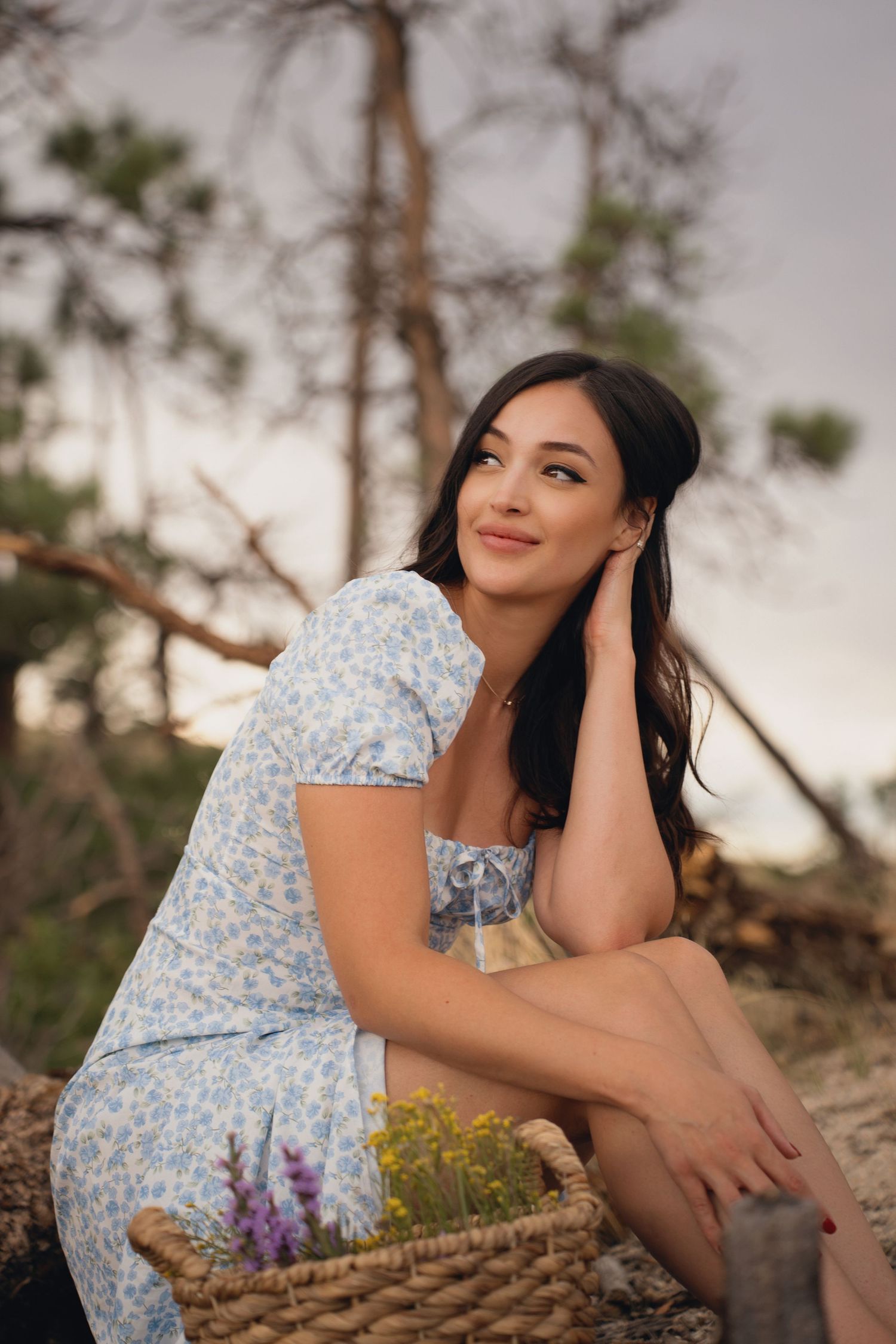 Woman in floral dress sitting outdoors with basket and wildflowers.