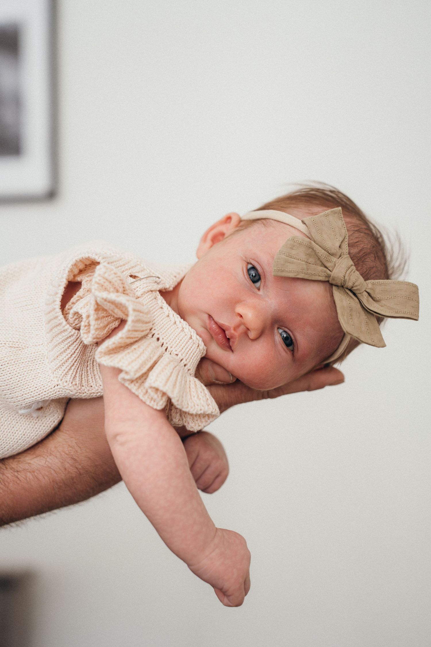 A baby wearing a cream knit outfit and tan bow headband rests gently in someone's arms.