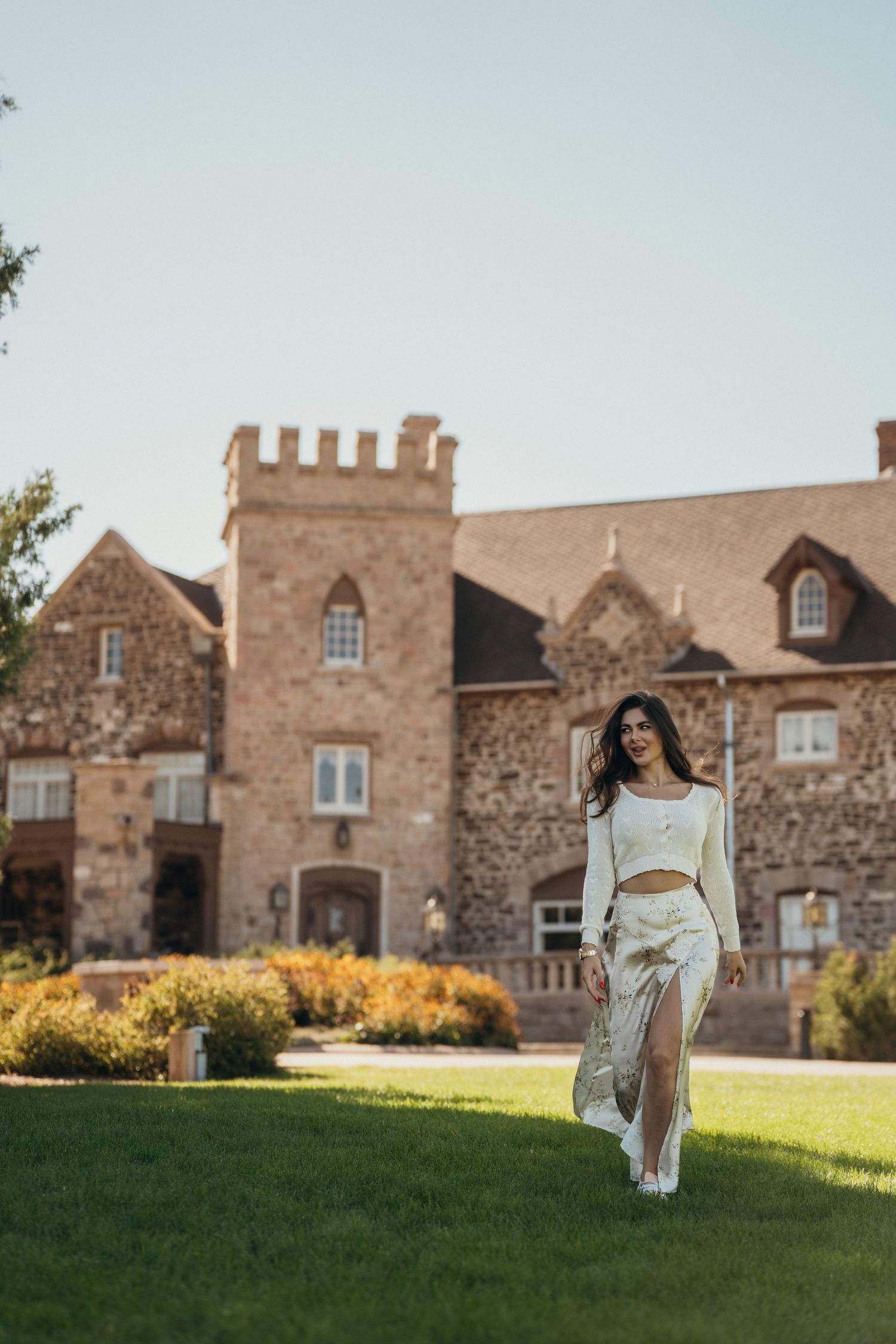 A person in white dress walks on a grassy hill in front of a stone castle-like building with turrets and windows.