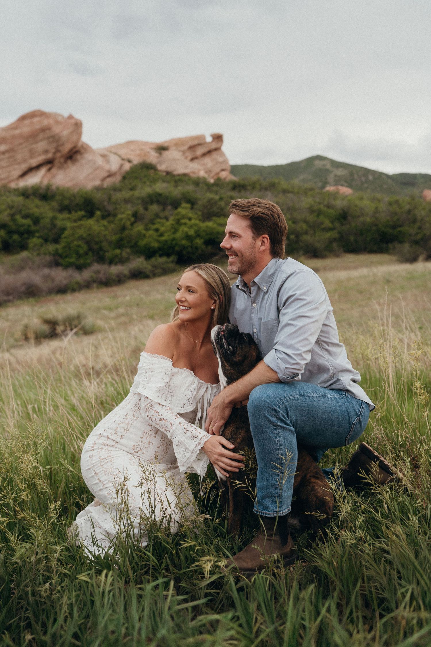 Couple smiling with their dog in a green meadow with red rock formations and mountains in the background.