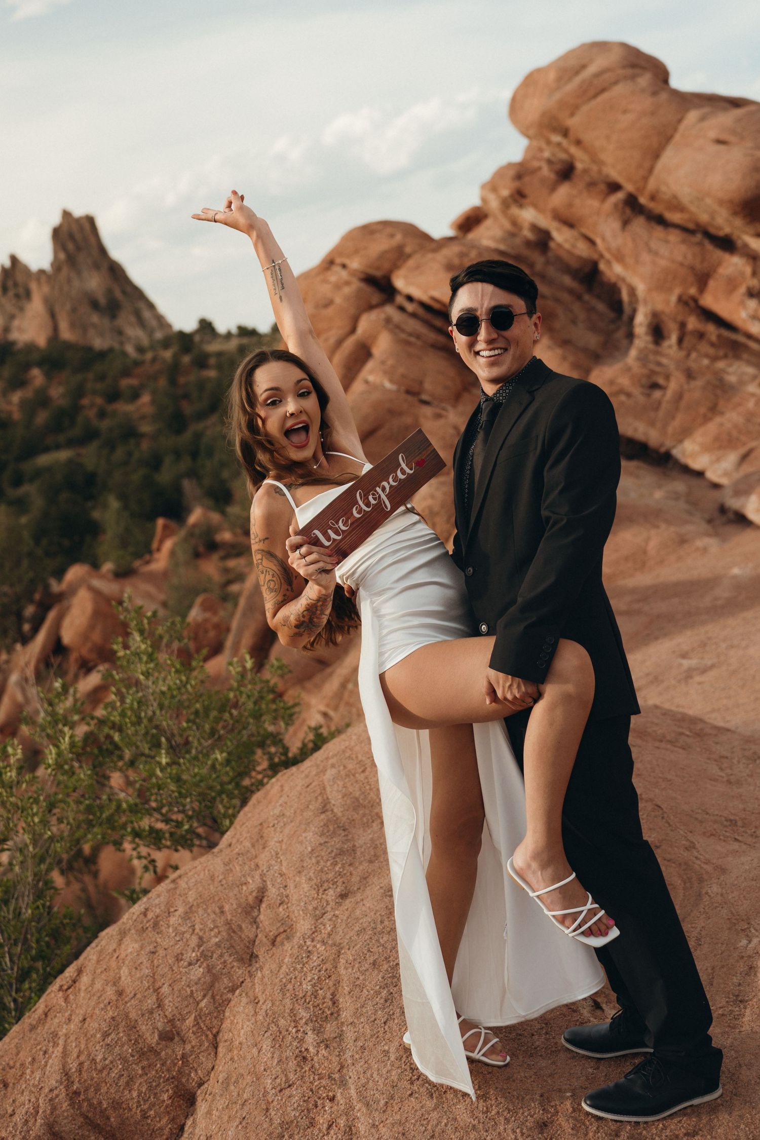 A couple in formal attire poses playfully on red rocks at sunset in an outdoor engagement photoshoot.