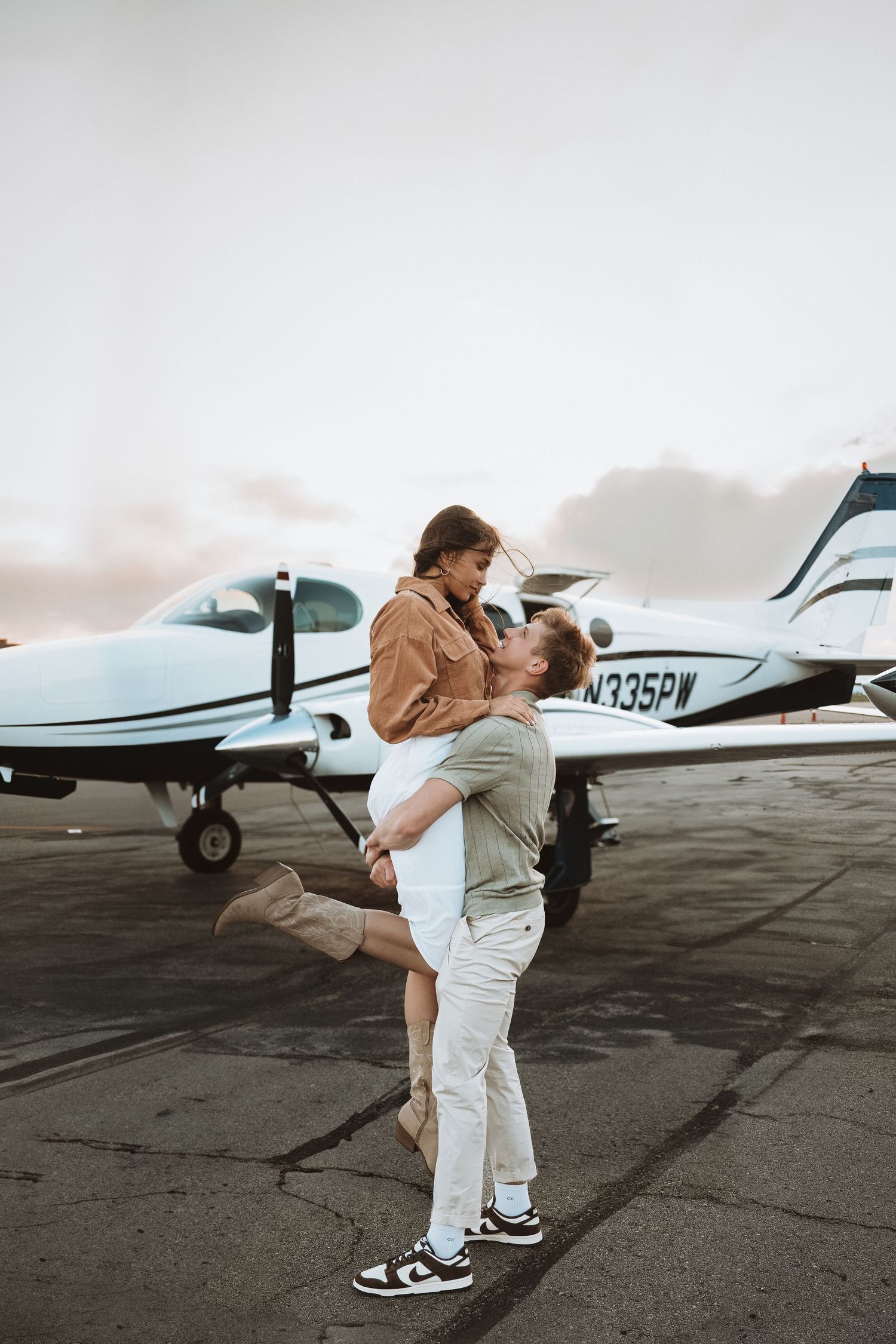 Romantic couple embraces on an airport tarmac with a small private airplane in the background during sunset.