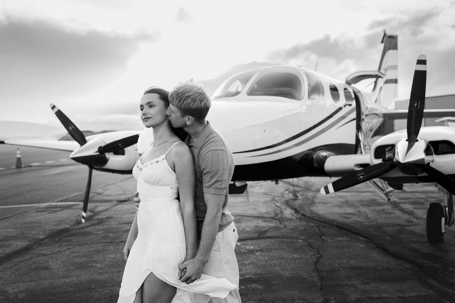 A romantic couple stands in an embrace in front of a small private airplane on an airport tarmac.