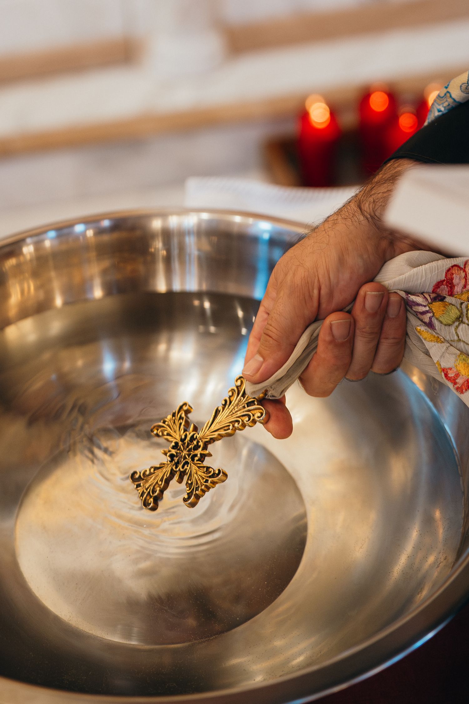 Elderly priest dipping ornate golden cross into silver baptismal font during religious ceremony.