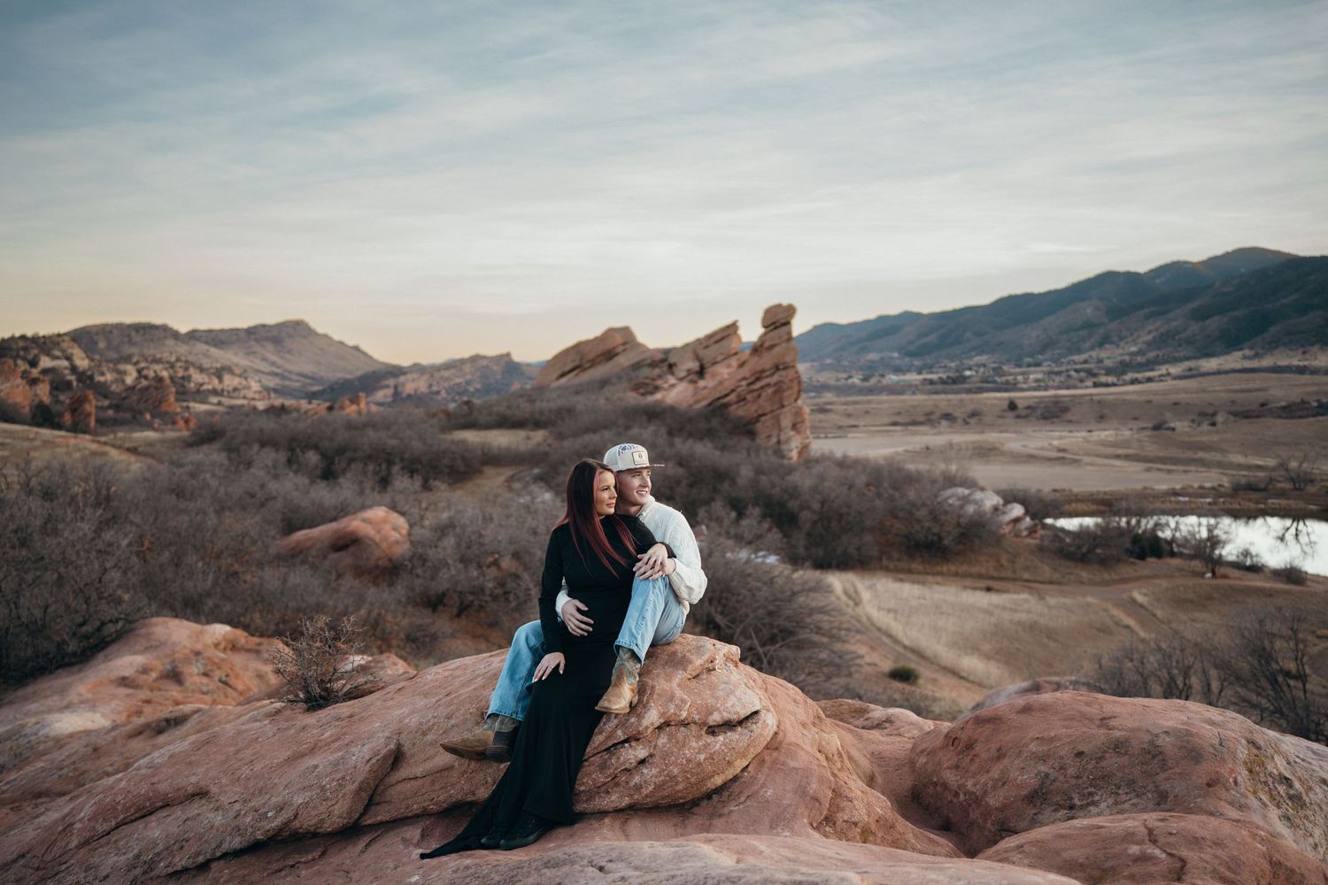 Two people embrace while sitting on red rocks overlooking a scenic mountain landscape at Red Rocks Park in Colorado.