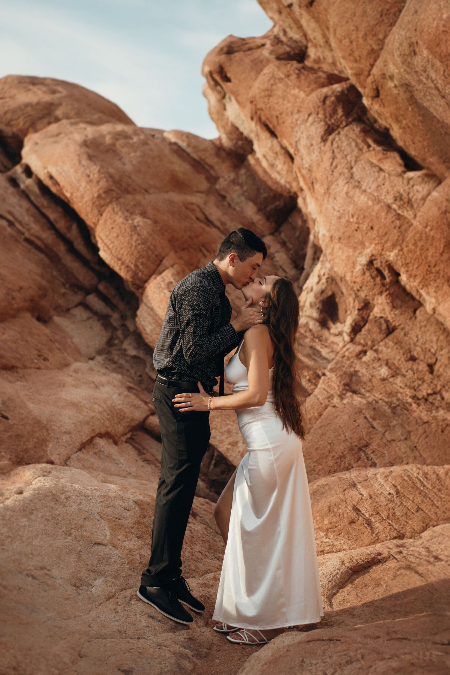 A romantic moment shared between a couple in elegant attire against dramatic red rock formations in a desert landscape.