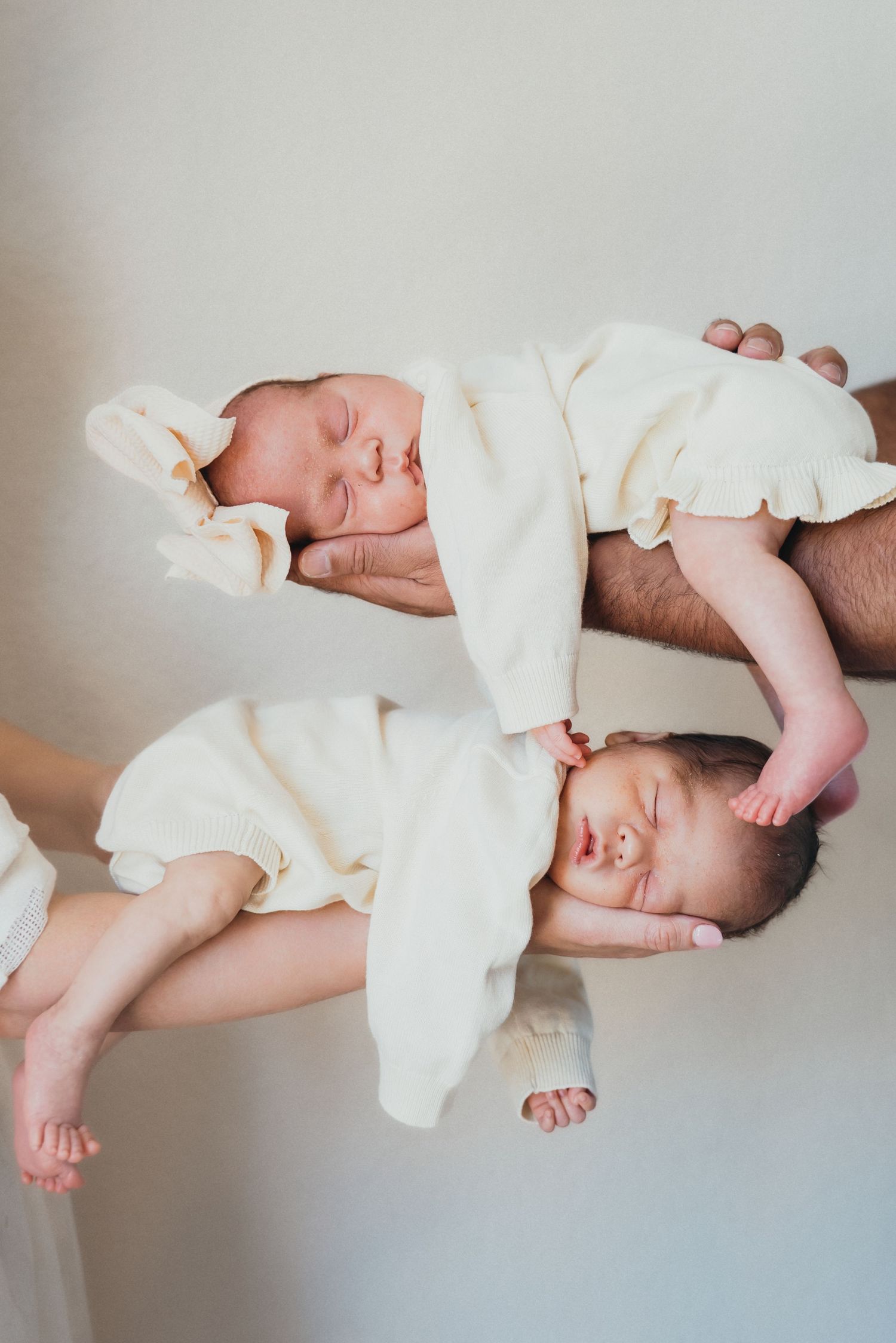 Two sleeping newborn twins in white onesies resting on top of each other against a light background.