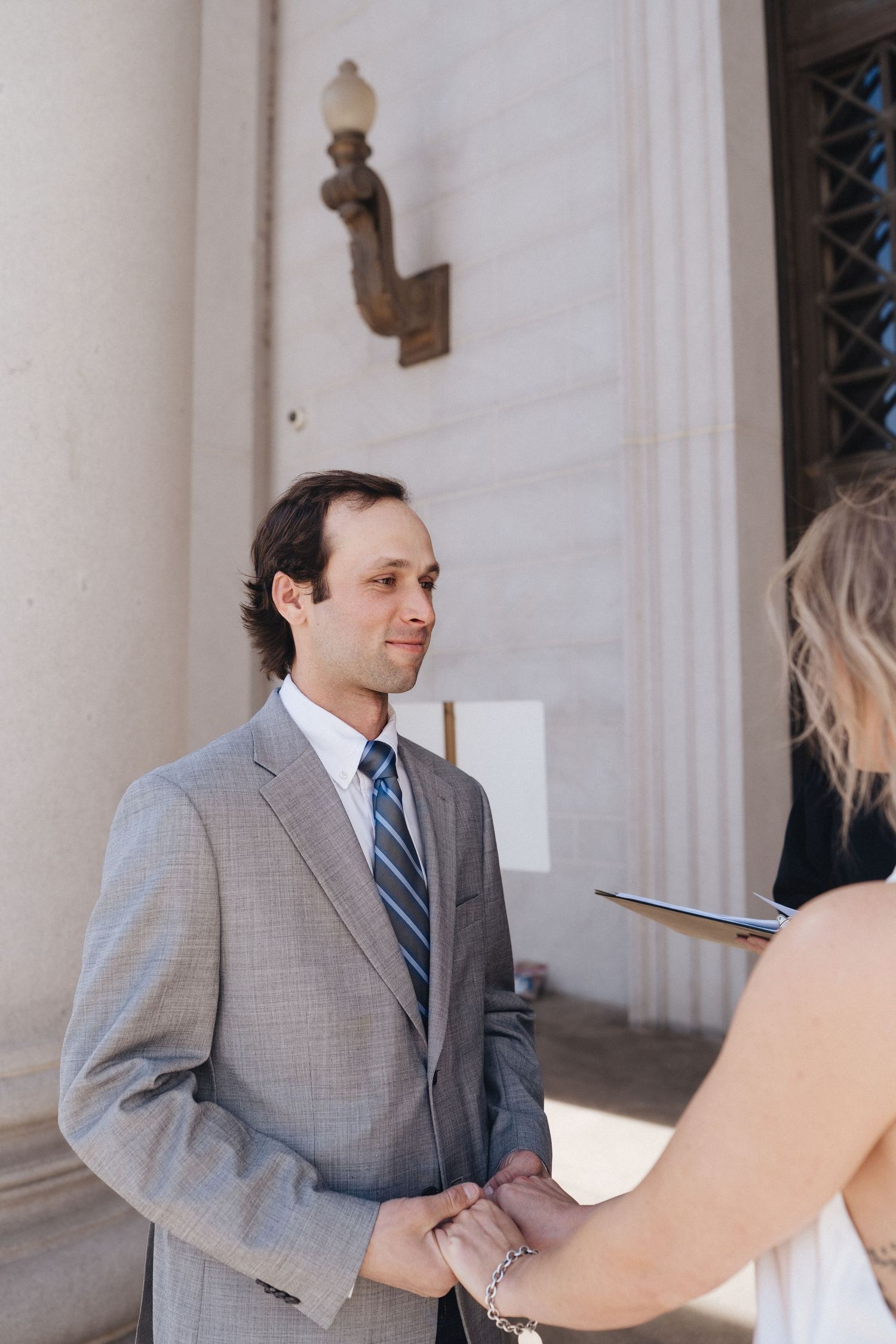 A person in a gray suit and striped tie stands in front of white columns at a formal building entrance.