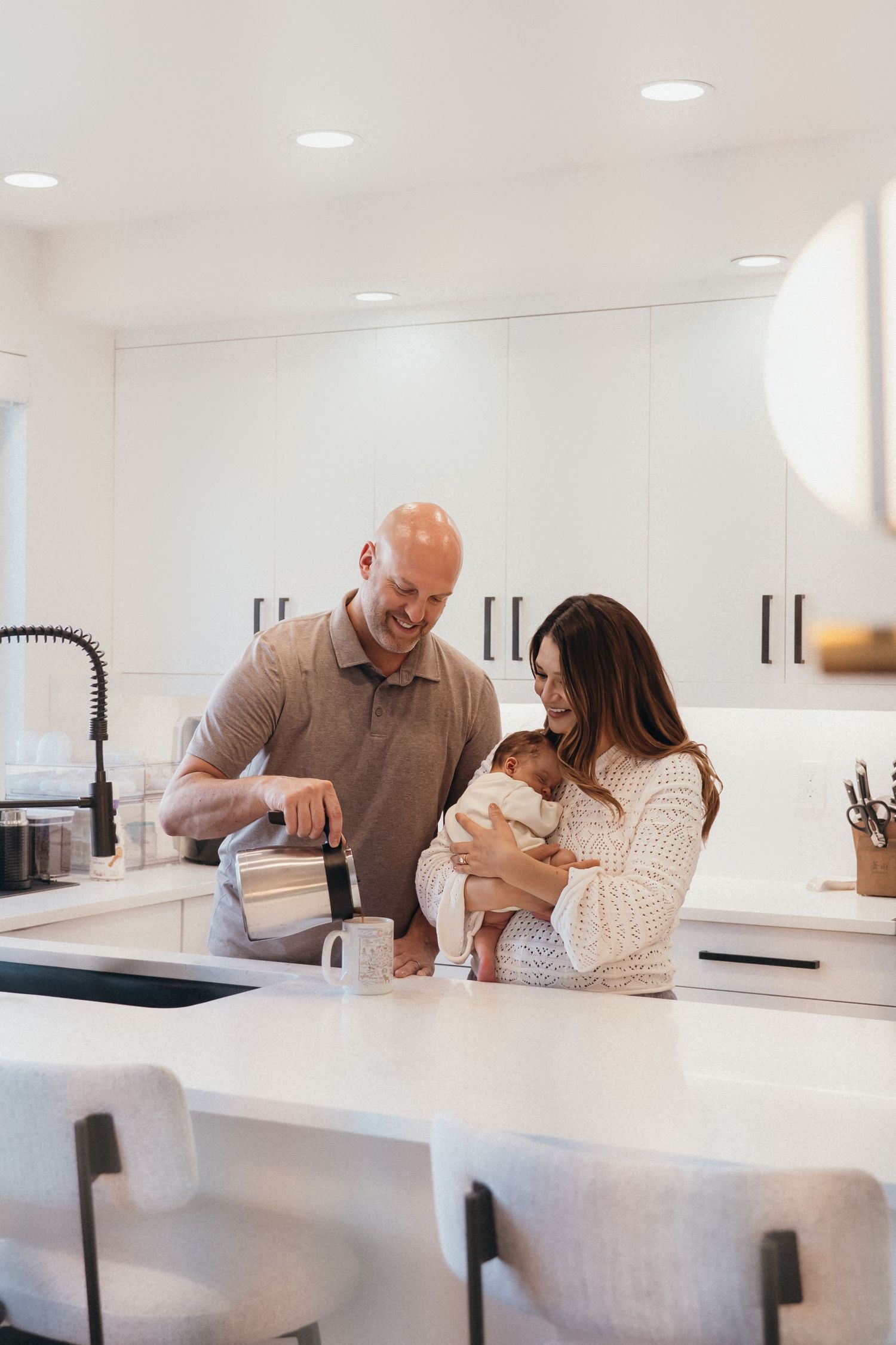 Family spending time together in modern white kitchen while preparing drinks and holding baby.