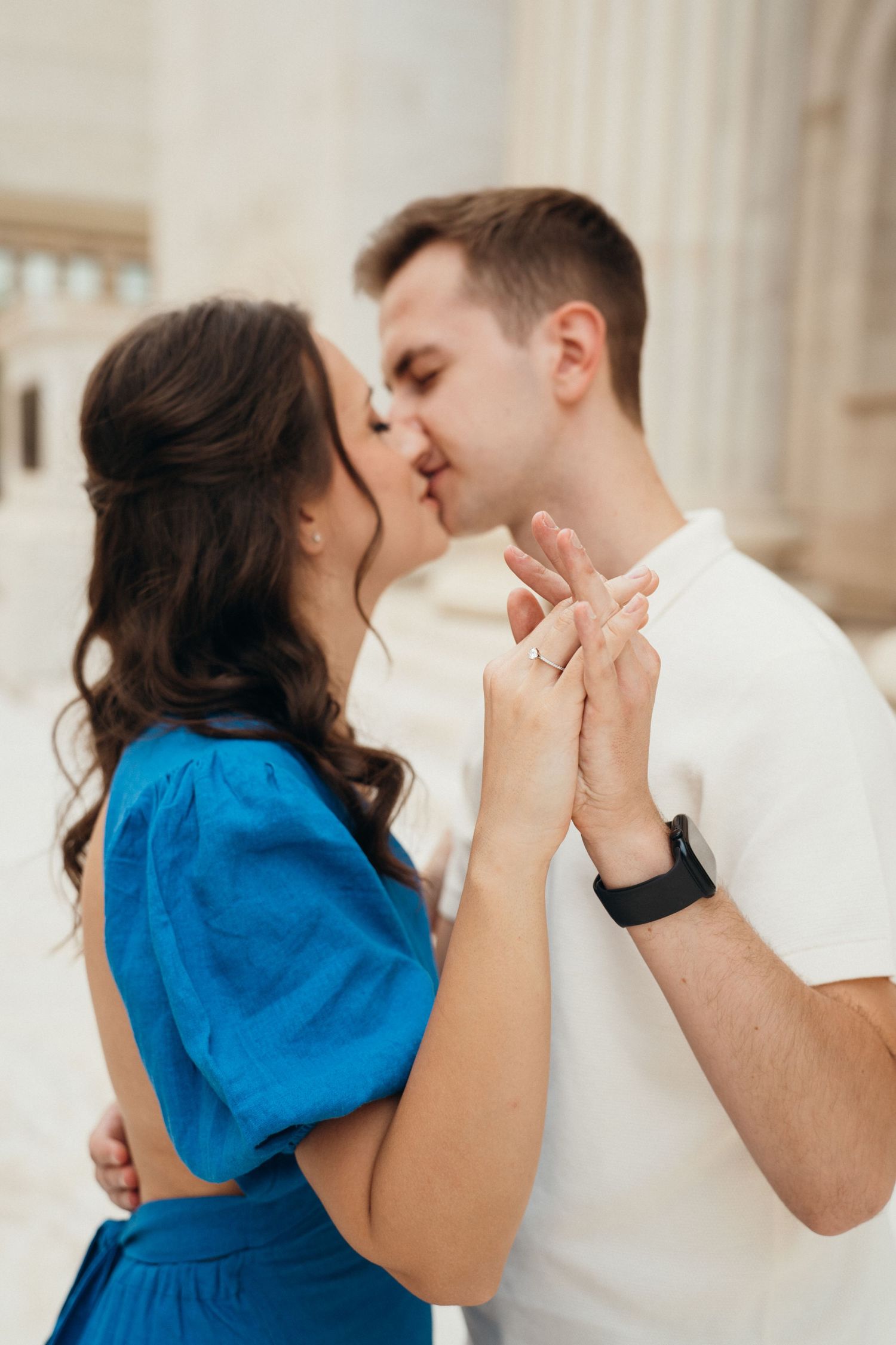 A romantic couple shares a tender kiss while holding hands, she in a bright blue dress and he in a white shirt.