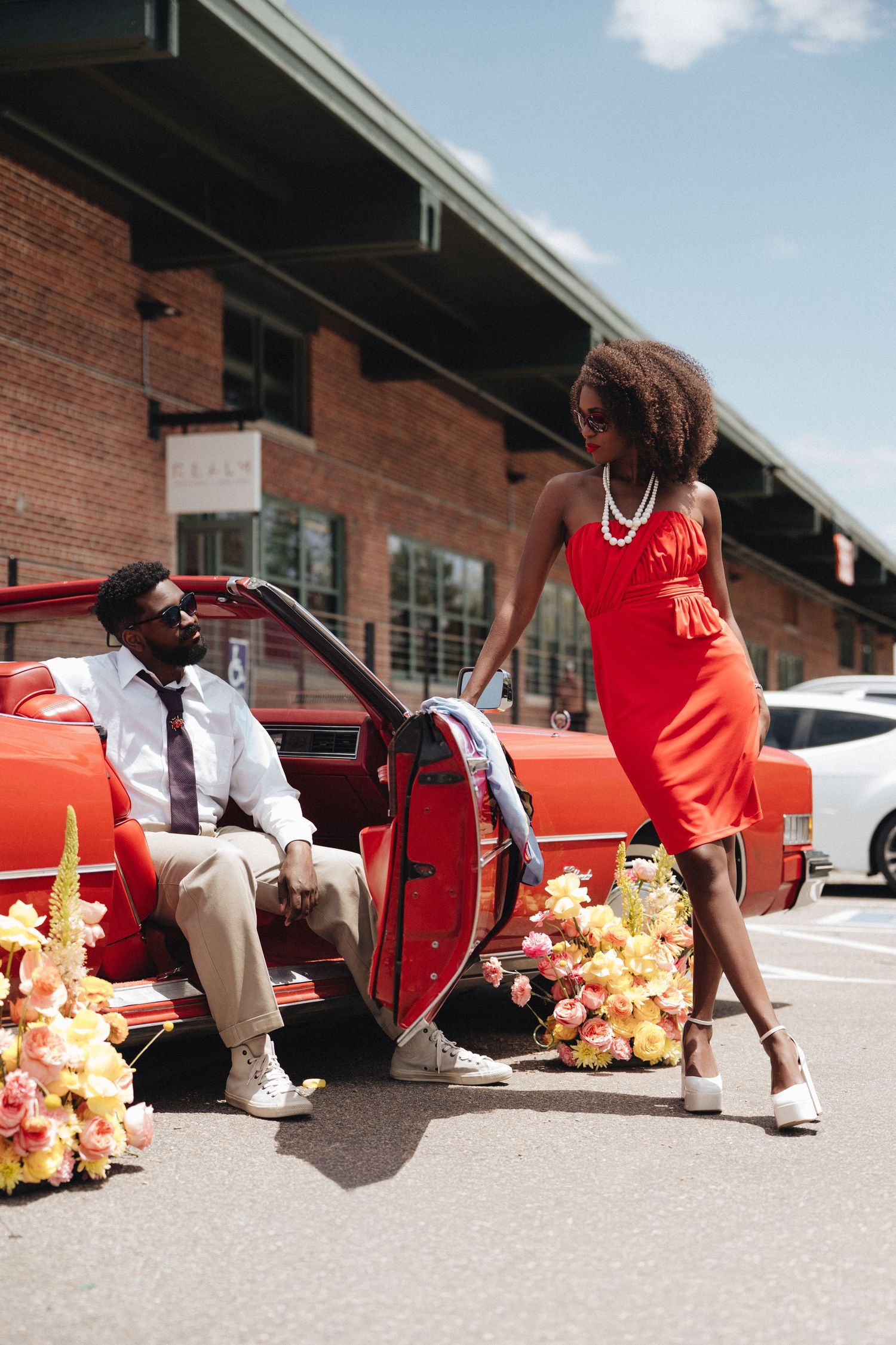A stylish couple poses beside a vintage red convertible adorned with flowers, woman in red dress, man in white shirt and tie.