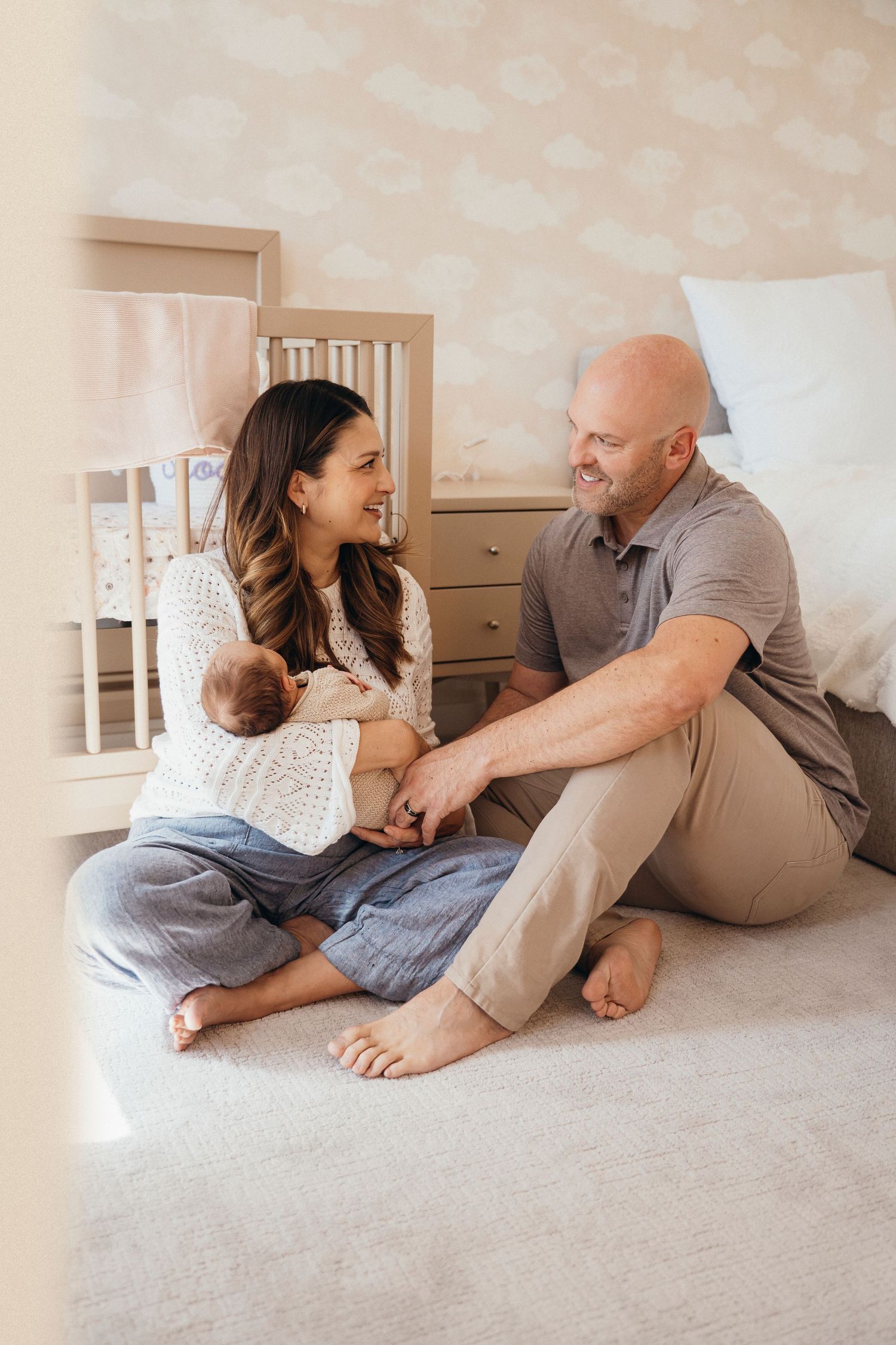 Family sits together on floor in nursery laughing and playing with infant wearing white sweater.