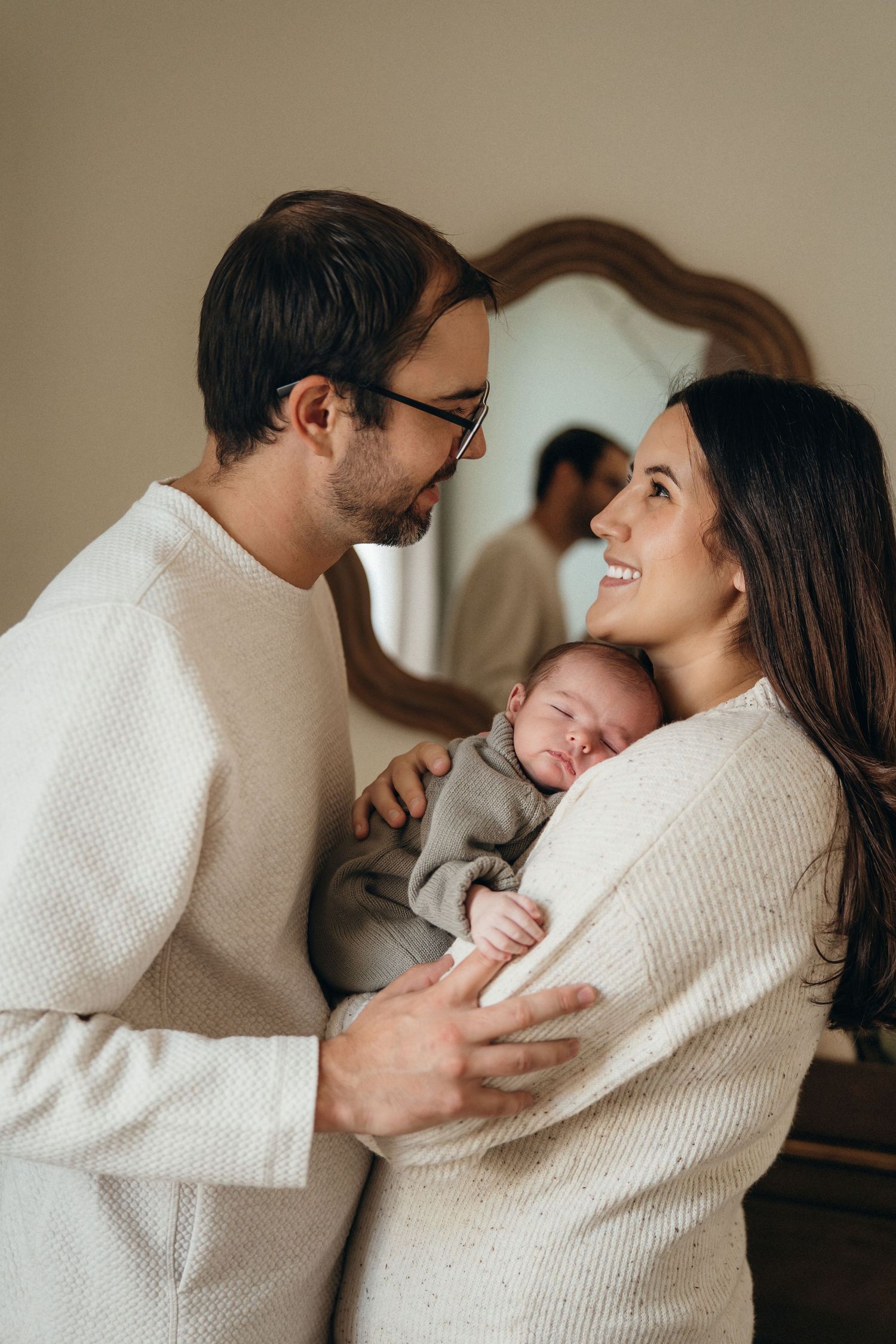 A couple in matching cream sweaters share a tender moment while holding their sleeping newborn baby.