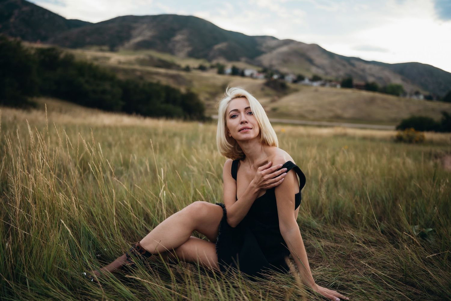 A person in a black dress poses gracefully in tall grass with rolling hills and mountains in the background.