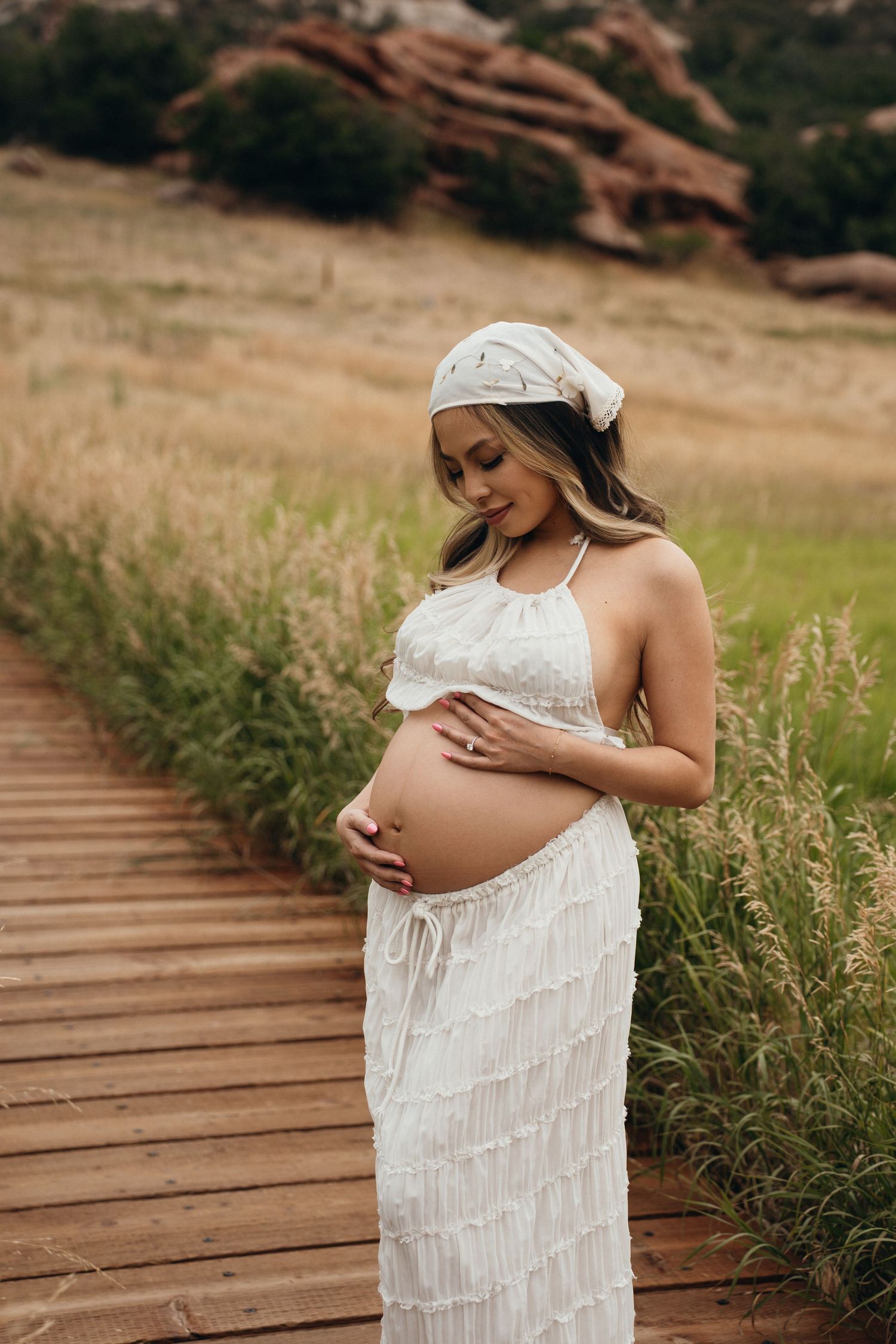A pregnant woman in a white fringe dress poses on a wooden pathway surrounded by tall grass in a natural outdoor setting.