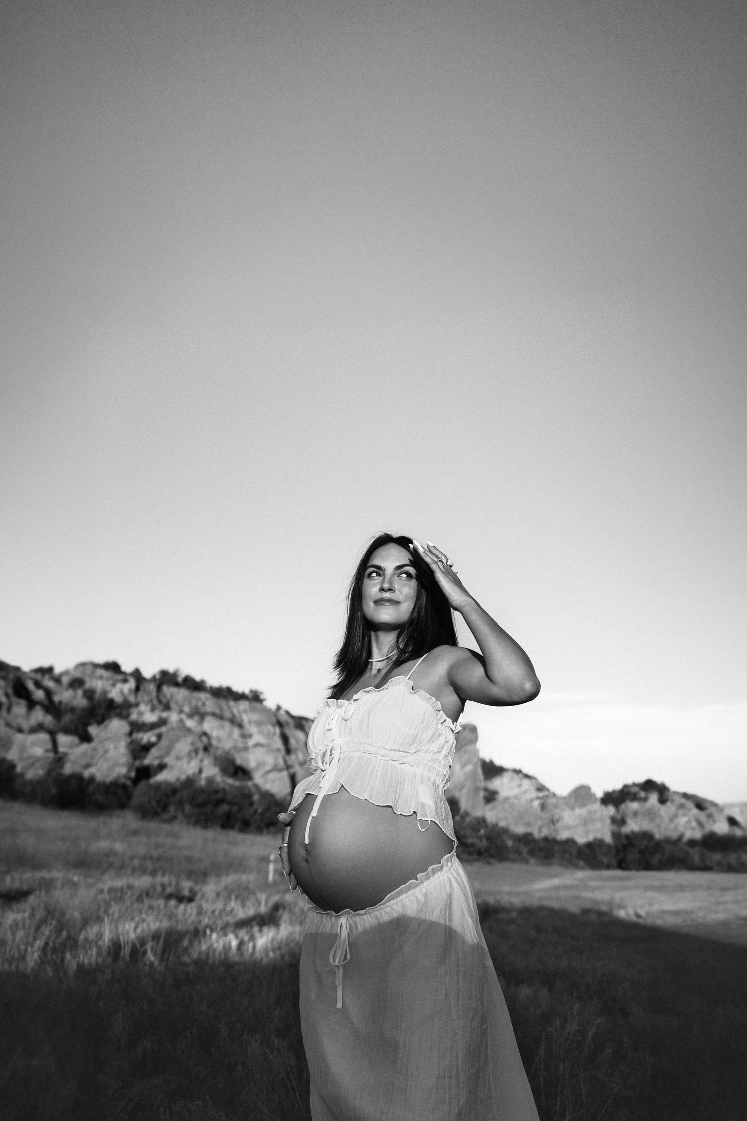 Black and white outdoor maternity photo showing a pregnant woman in flowing dress standing in a natural landscape.