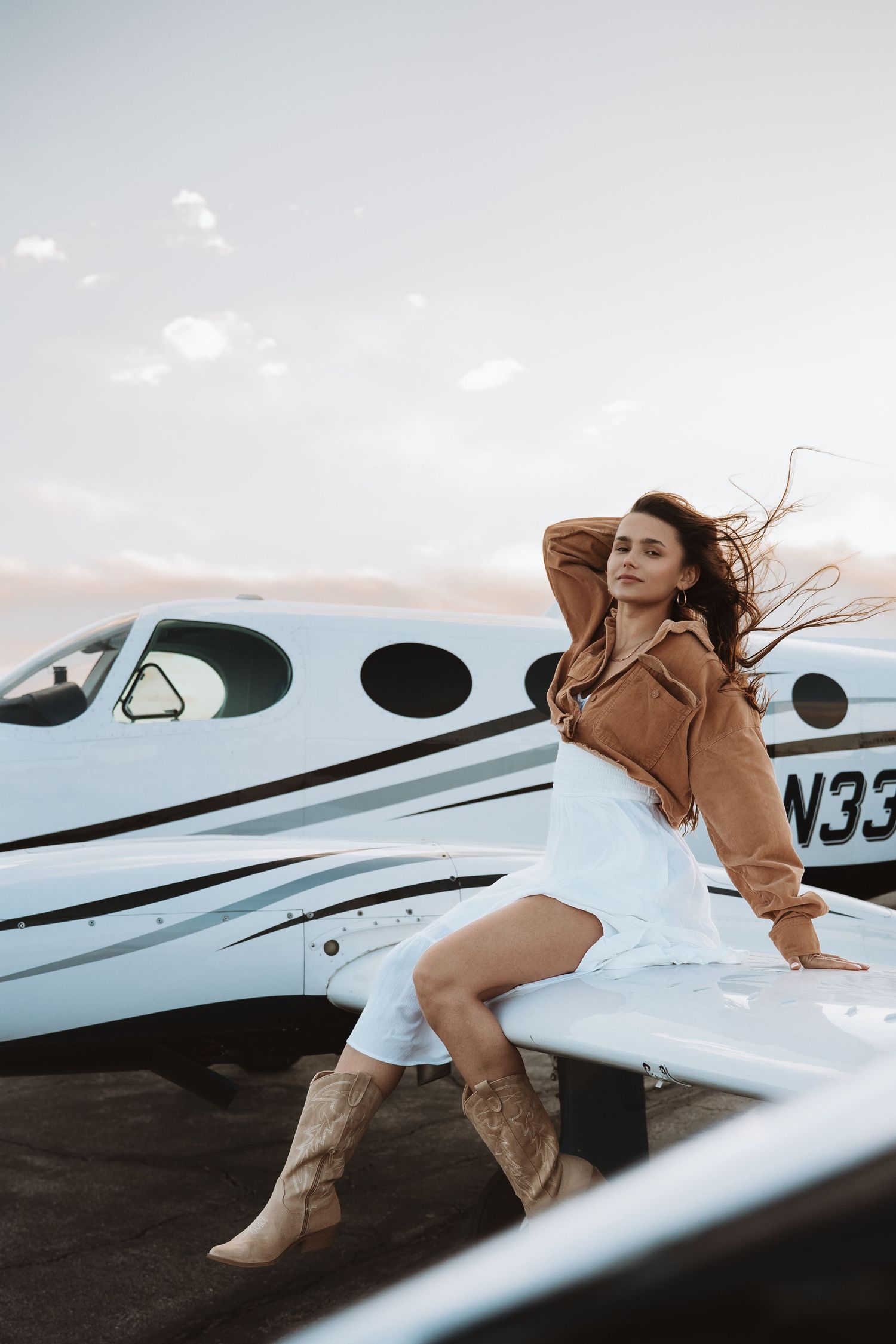 Fashion photoshoot featuring a brown jacket and boots pose on a small private aircraft wing at sunset.