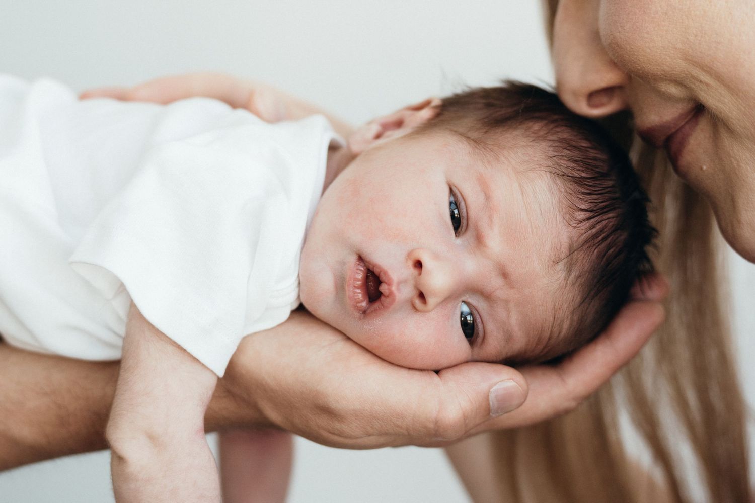Baby in white clothing rests in gentle hands while looking up with bright eyes and an open expression.