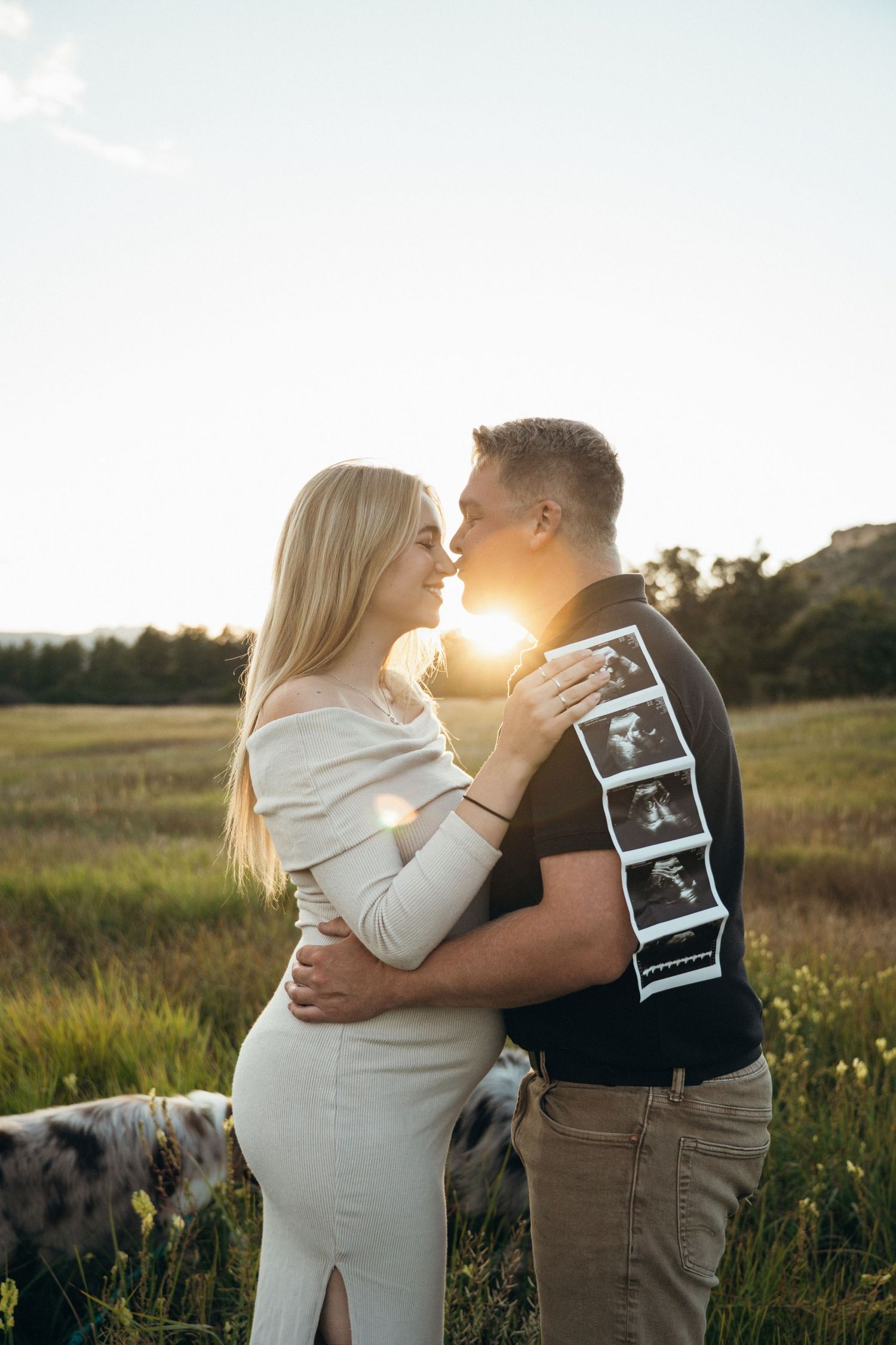 A couple embraces in a sunlit field during a pregnancy announcement photoshoot with an ultrasound photo.