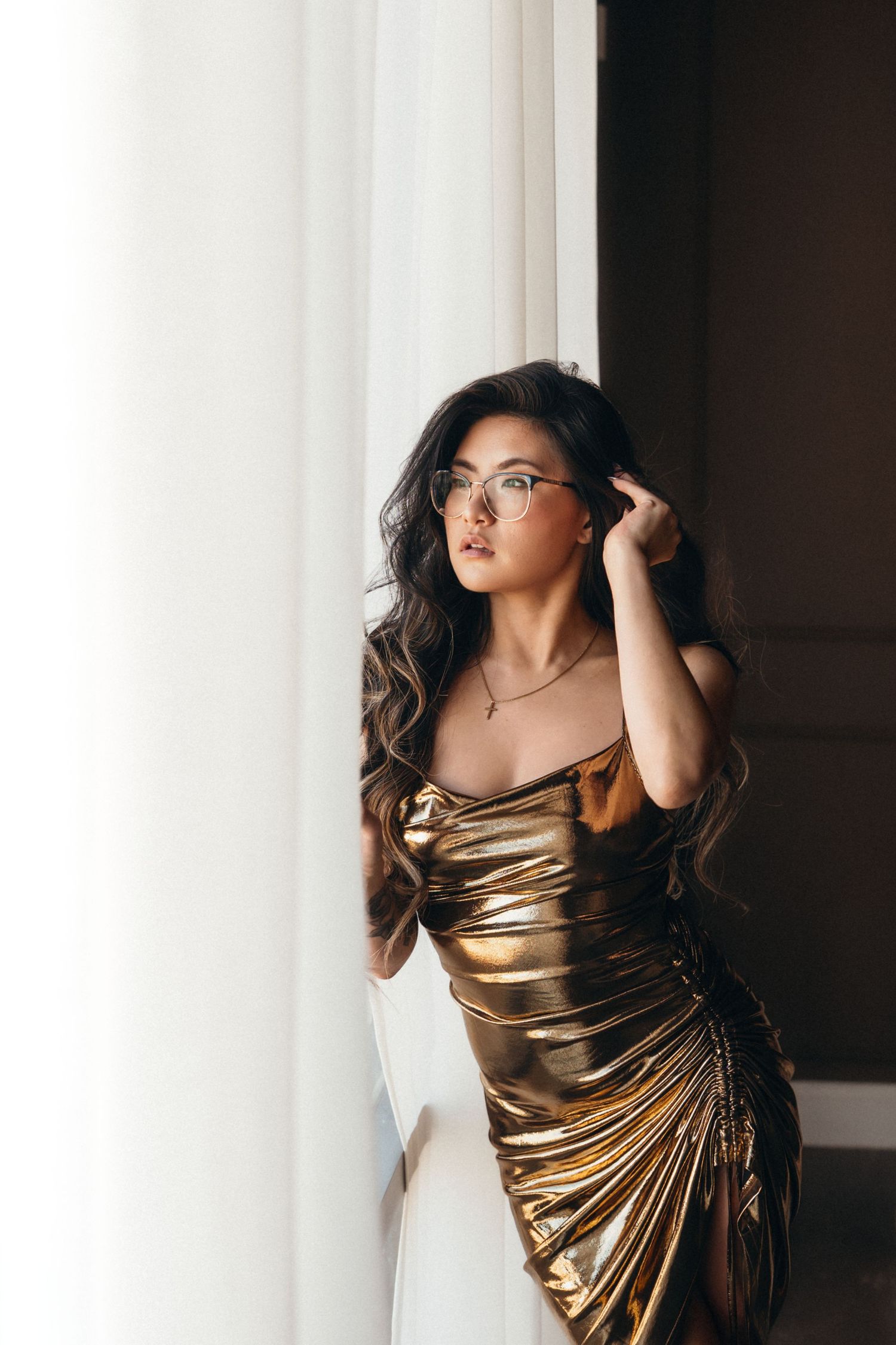 Glamorous woman in metallic gold dress with glasses poses elegantly by window with white curtains.