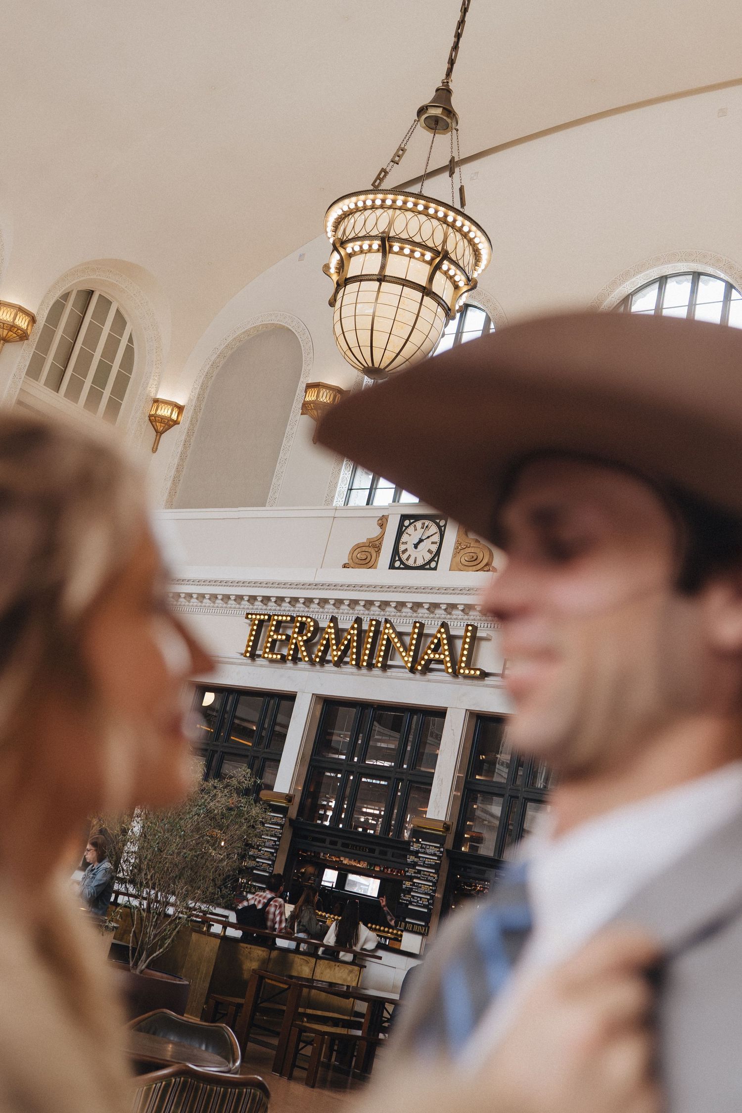 Motion blurred view of people in western attire inside historic train station with elegant chandeliers and arched windows.