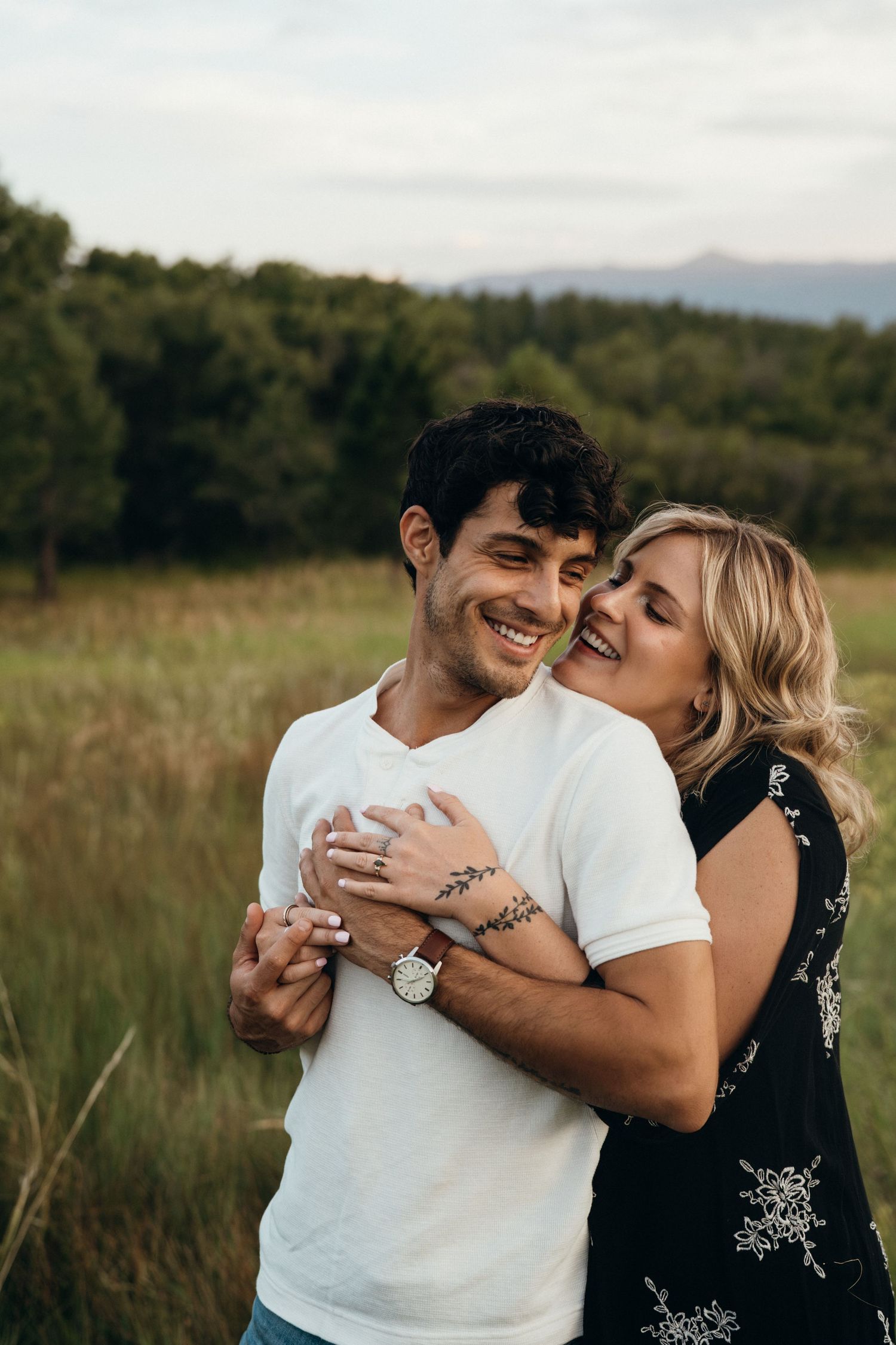 A happy couple embraces in a grassy field with mountains visible in the background during golden hour.