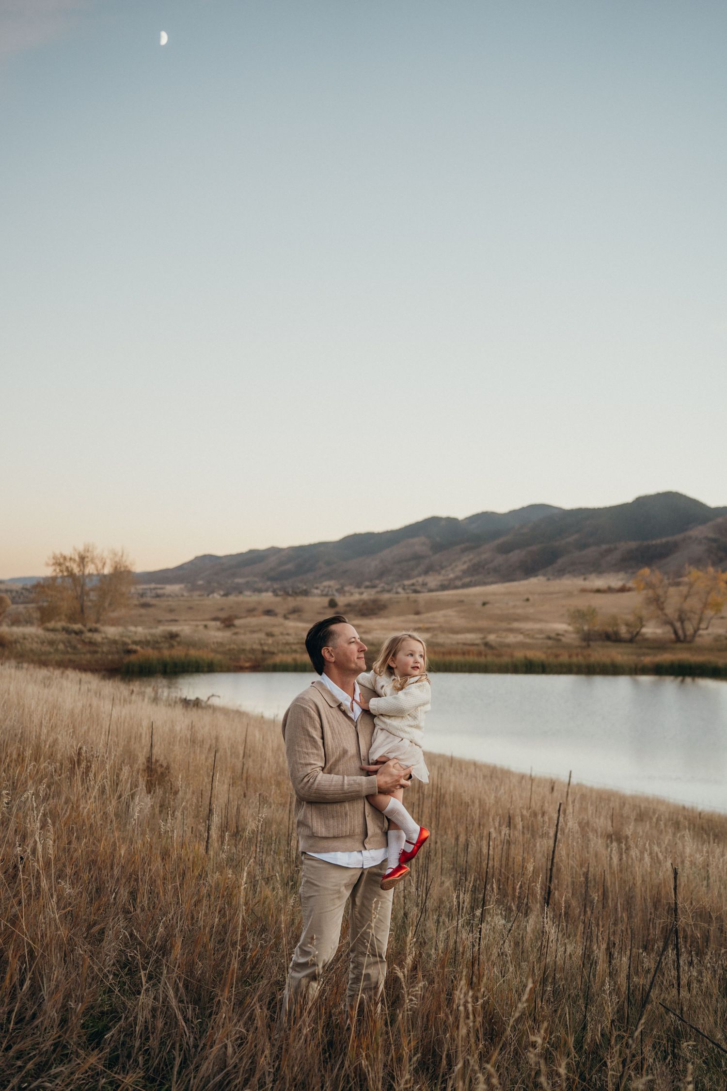 Scenic lakeside landscape with tall grass and mountains at sunset in warm golden light.